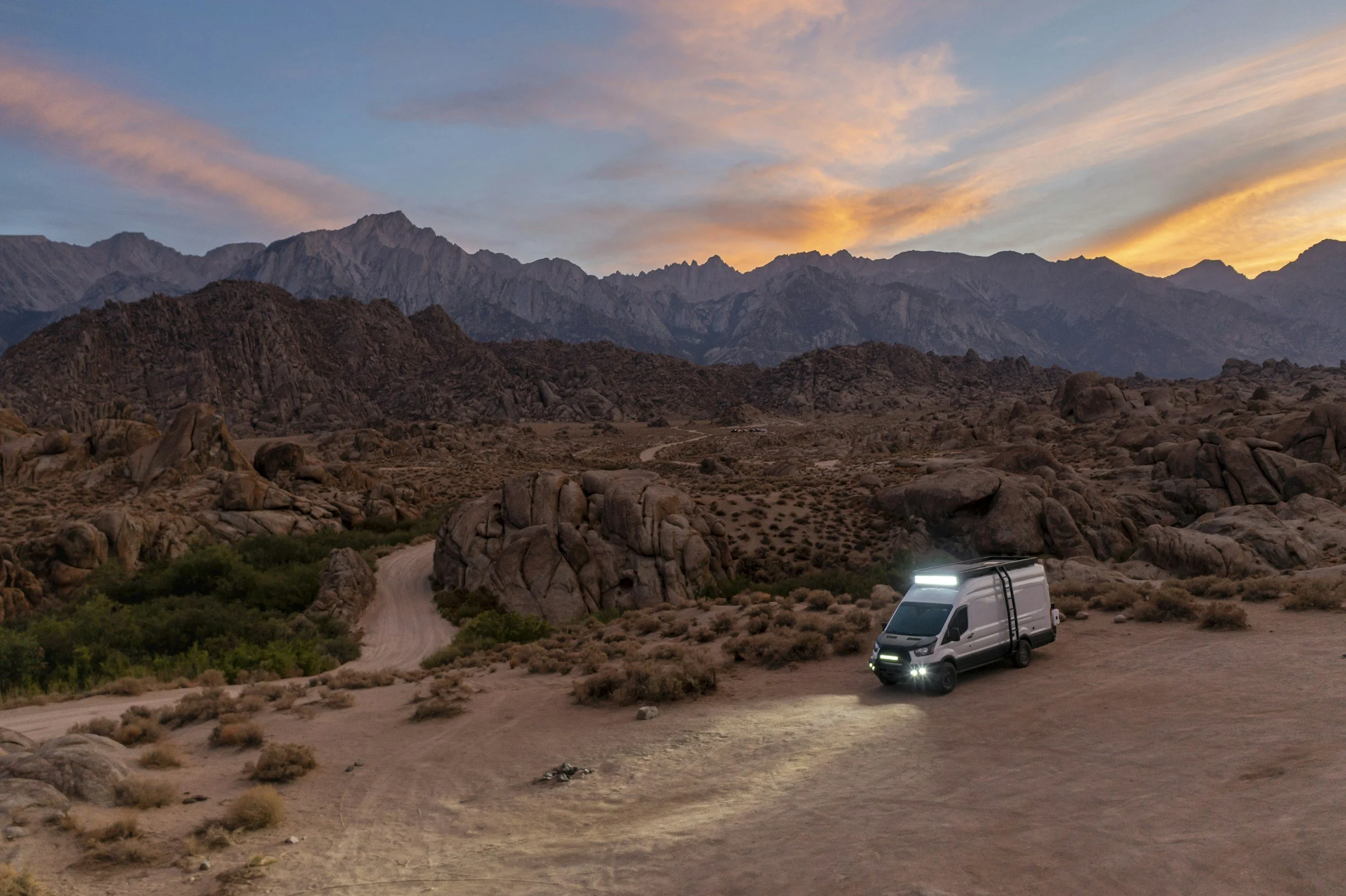 A white van with bright headlights parked on a dirt track in a desert landscape at sunset, with rocky hills and mountains in the background.