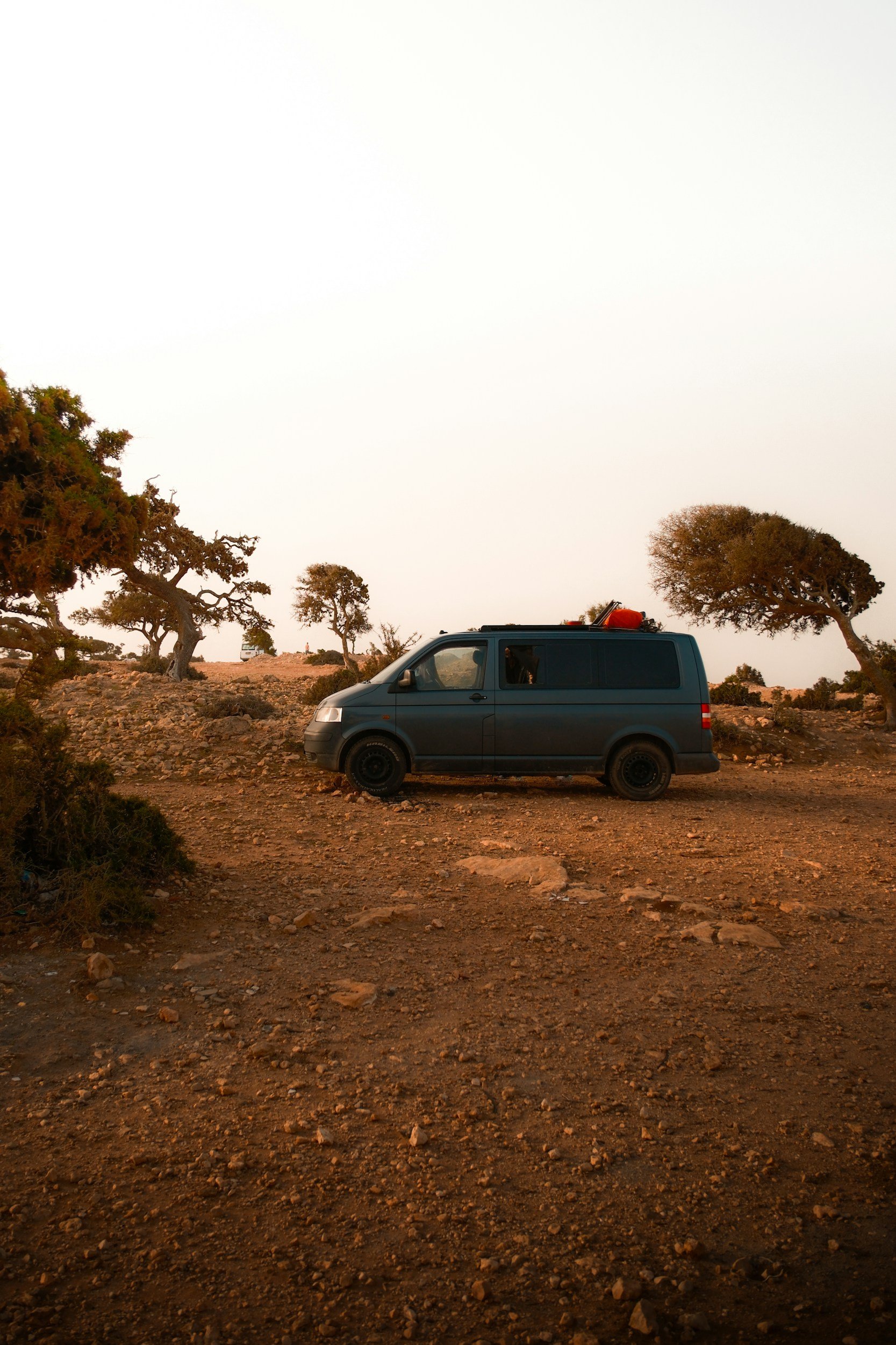 A dark blue van parked on a dirt trail in a desert landscape with sparse trees, under a pale sky at sunset.