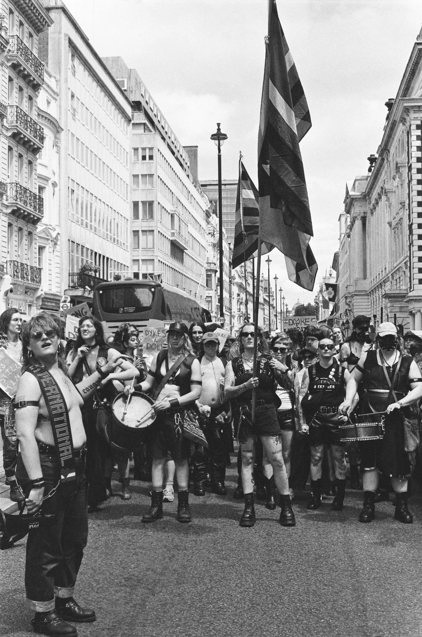 Black and white photo of a street protest with many protesters holding flags and signs. People are dressed in alternative and punk fashion styles, some with tattoos, sunglasses, and accessories. Venue appears to be an urban area with tall buildings.