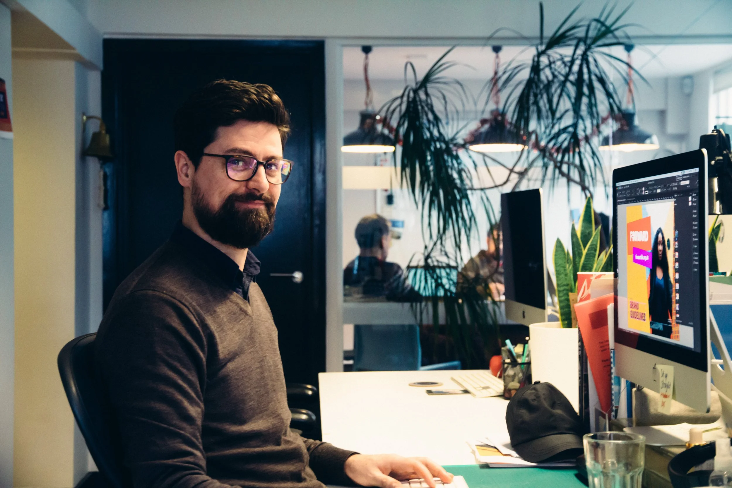 A man with glasses, dark hair, and a beard sitting at a desk in an office, working on a computer with colorful graphic design on the screen. There are plants, notebooks, and other office supplies on the desk.