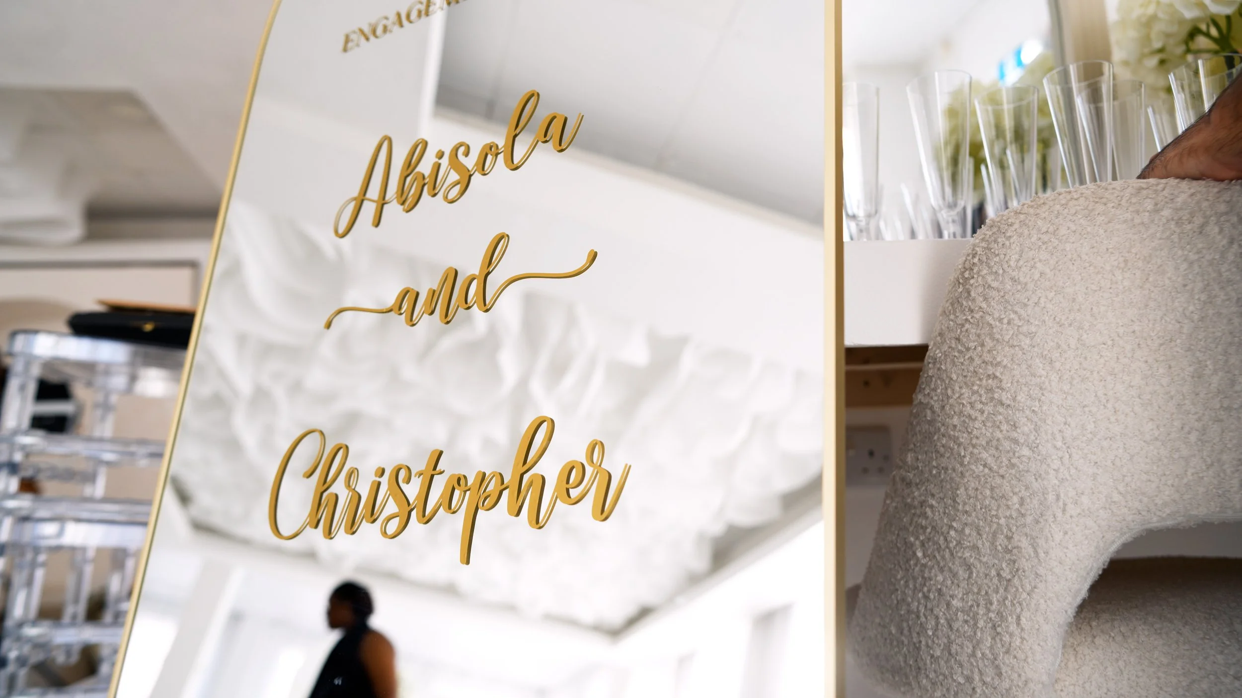 A gold-framed mirror with the text 'Abisola and Christopher' in gold cursive, reflecting a woman in black standing in a bright room with white stone walls and a woman in a beige textured fabric chair, surrounded by clear glassware.