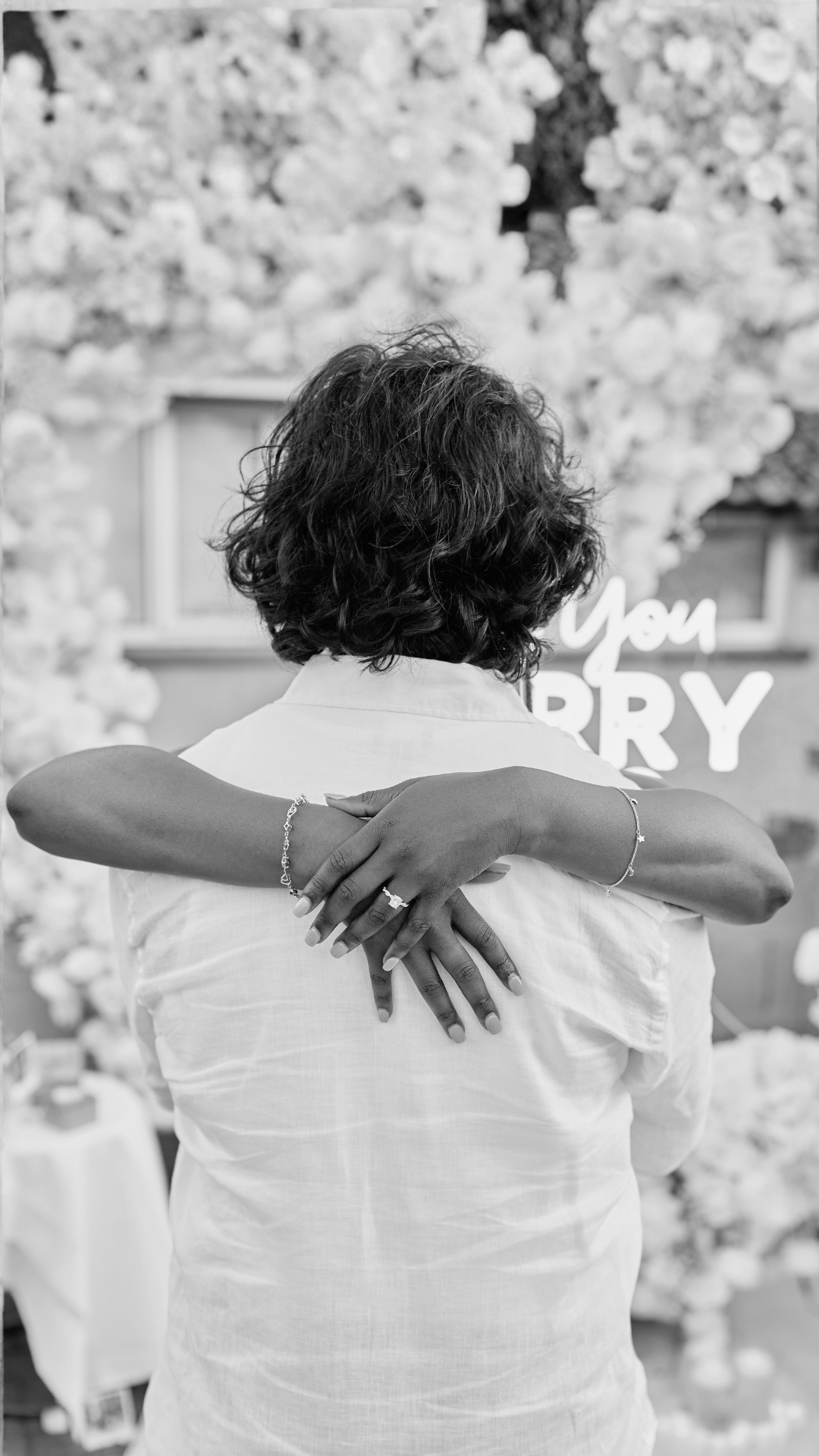 A woman with short, curly hair is hugging herself with crossed arms, wearing rings and bangles, with a blurred background of flowers and a sign partially visible reading "You Sorry."