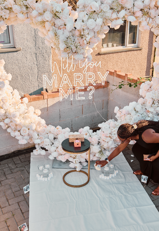 A woman arranges candles on a white cloth during an outdoor wedding proposal setup with a rose arch and a neon sign that reads "Will you MARRY ME?" in front of a house wall.