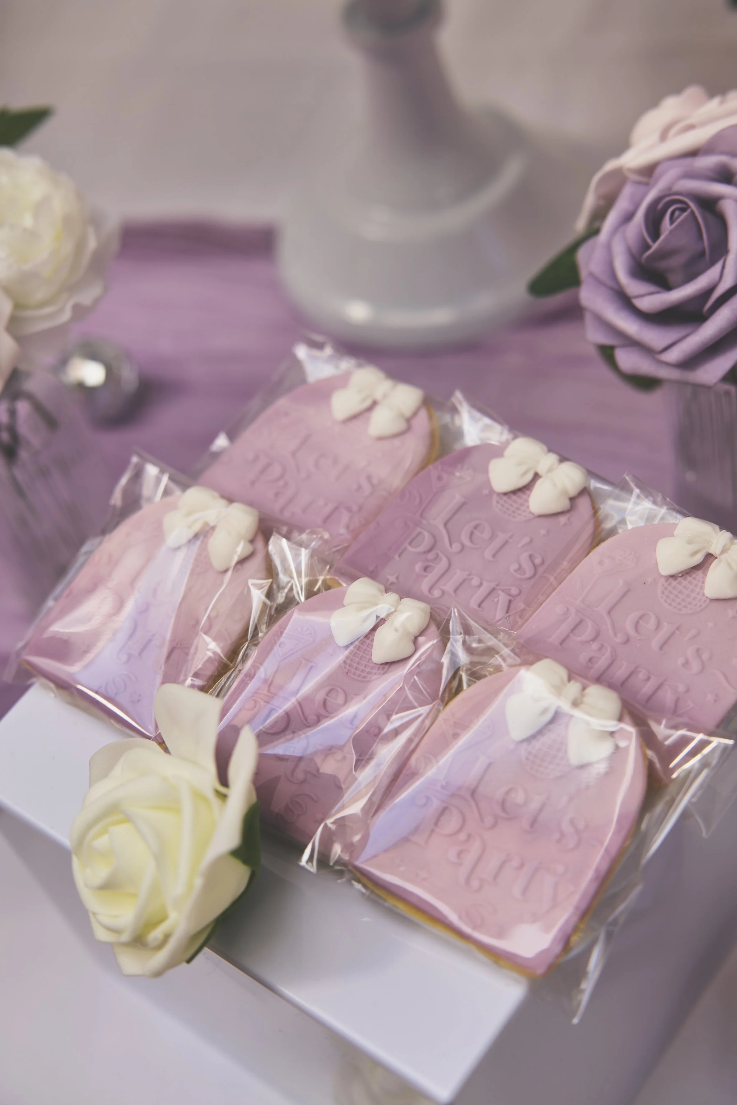 Pink custom cookies with heart decorations inside a clear plastic wrap, arranged on a white surface, surrounded by white and purple roses.