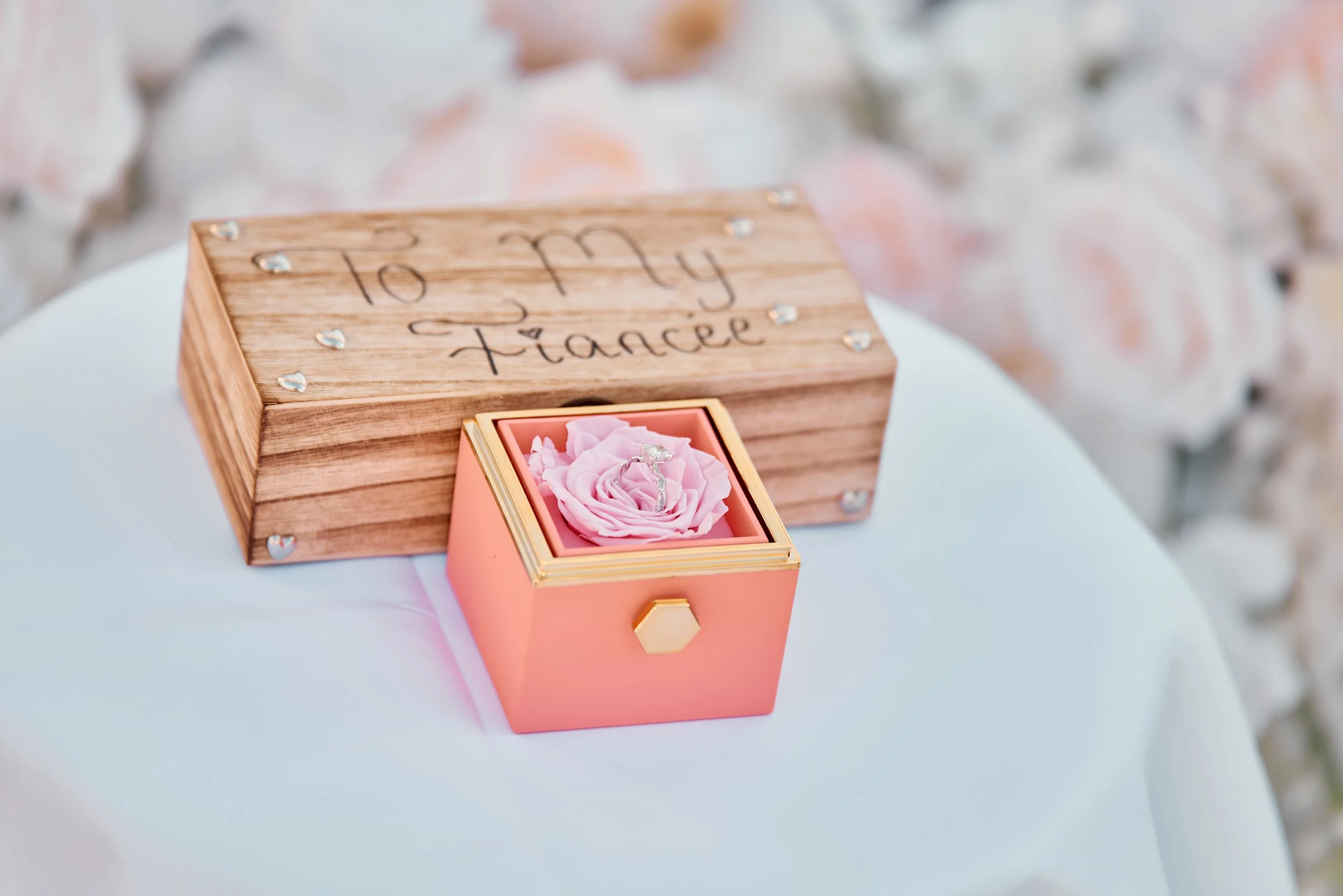 A wooden box with the words "To My Fiancée" written on it, placed on a white surface, with a small pink gift box featuring a clear ring inside on top of a pink rose.
