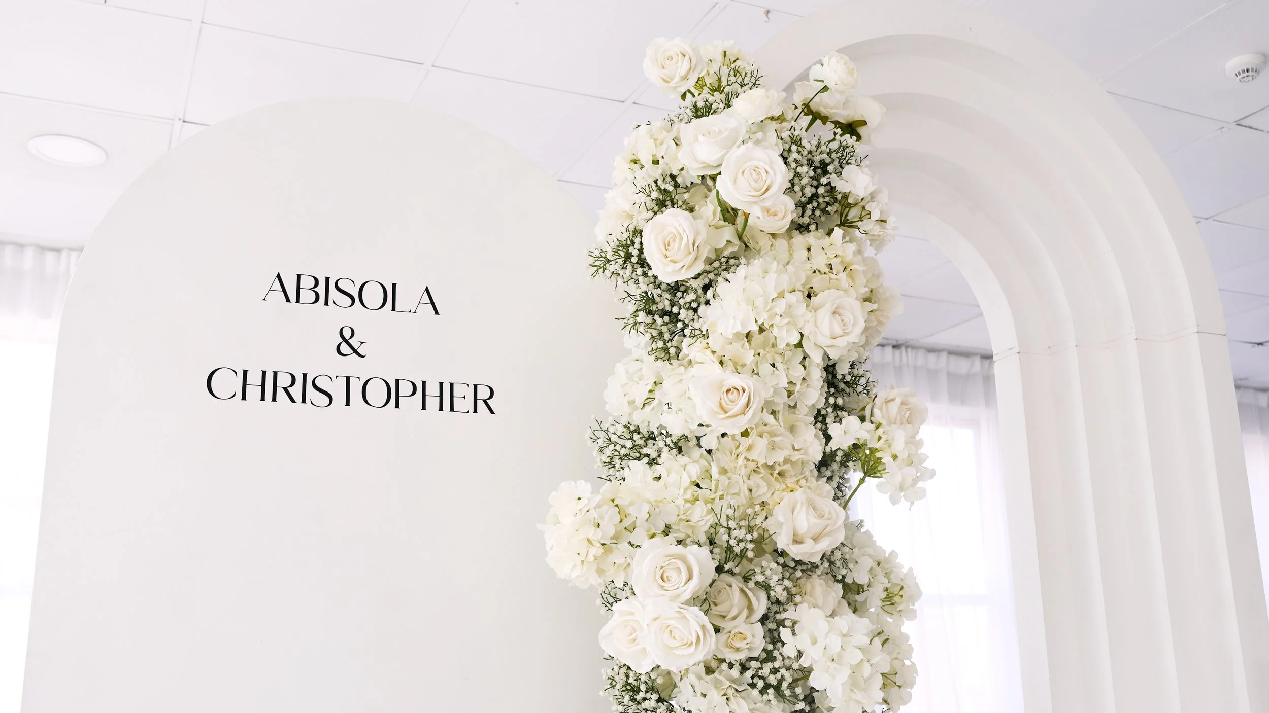 Wedding sign with black text reading 'Abisola & Christopher' on a white background, decorated with white roses and flowers on the right side, and set against a white indoor venue with curved architectural design.