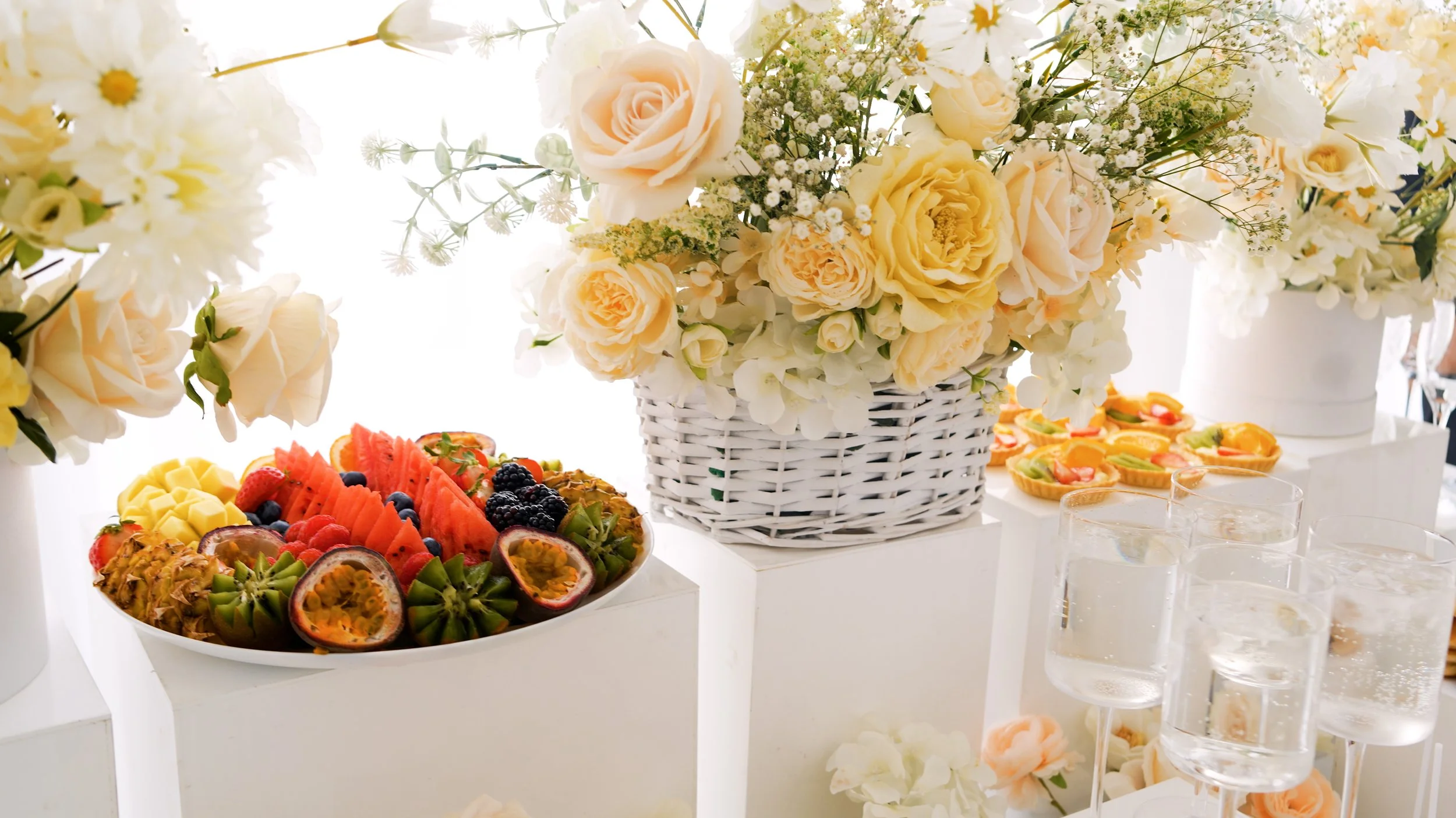 Elegant fruit platter with watermelon, pineapple, passion fruit, and berries surrounded by flower arrangements in white vases and glasses of water.