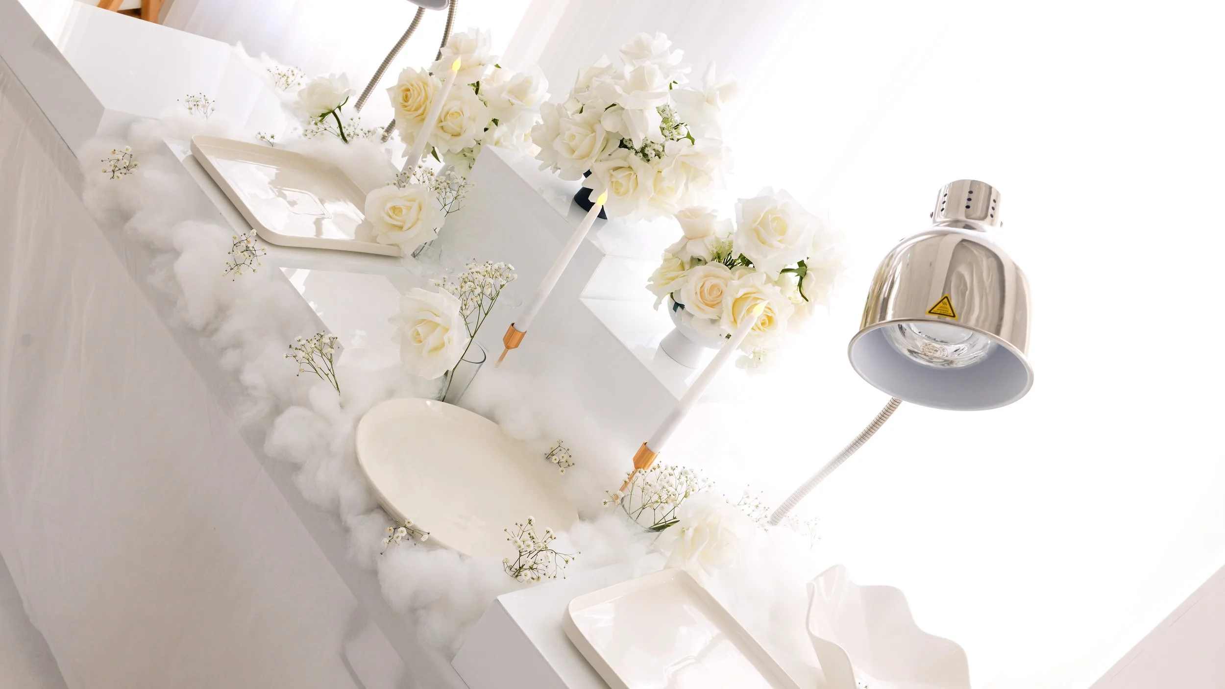 White table decorated with white roses, candles, and fluffy cotton, with a large silver lamp overhead.