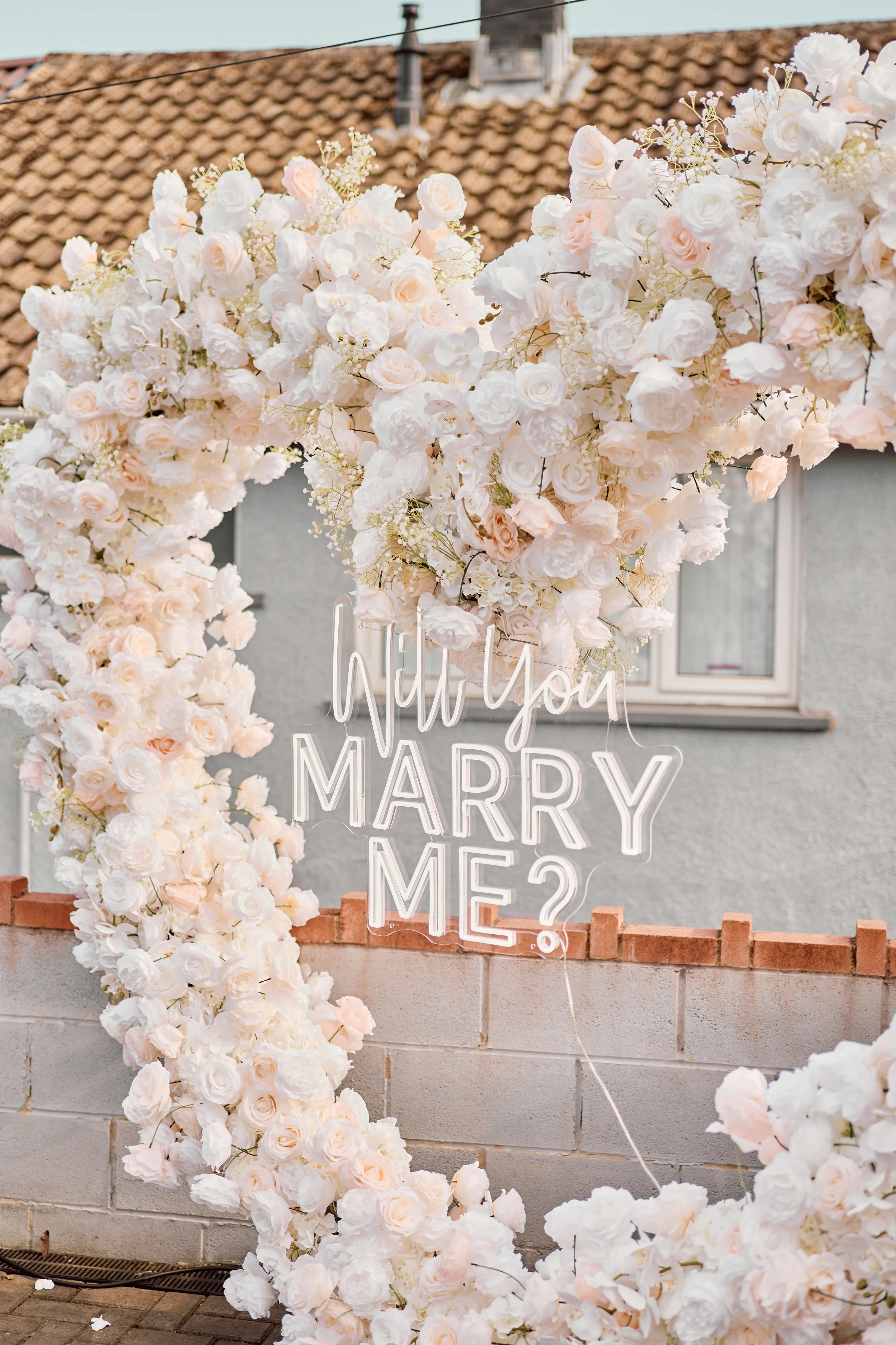 A wedding arch made of white and blush pink flowers with a neon sign that says 'Will you MARRY ME?' hanging among the flowers.
