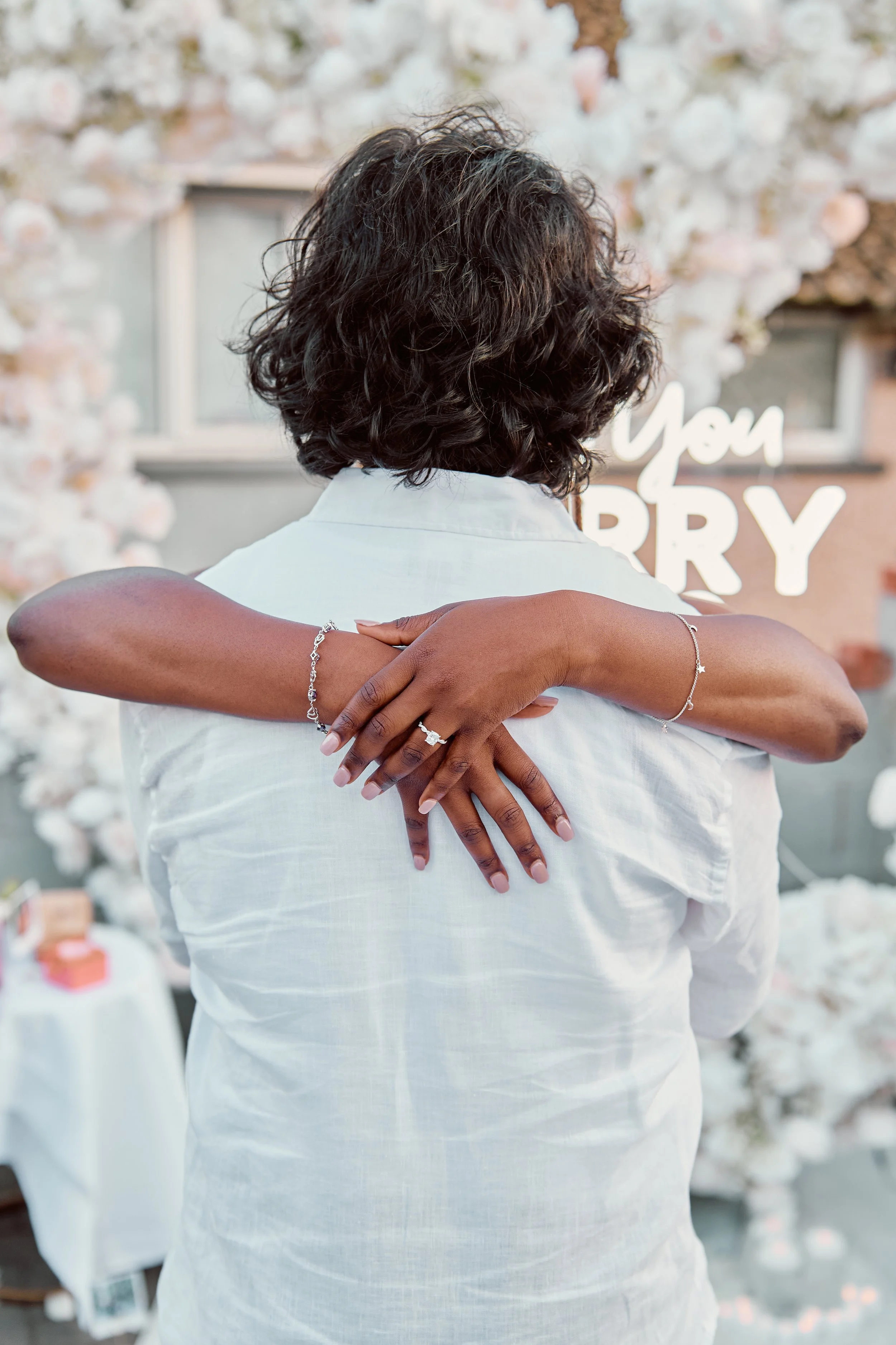 A person with curly black hair embracing another wearing a white shirt, showing hand jewelry, in front of a floral backdrop and a sign that says "You Sorry."