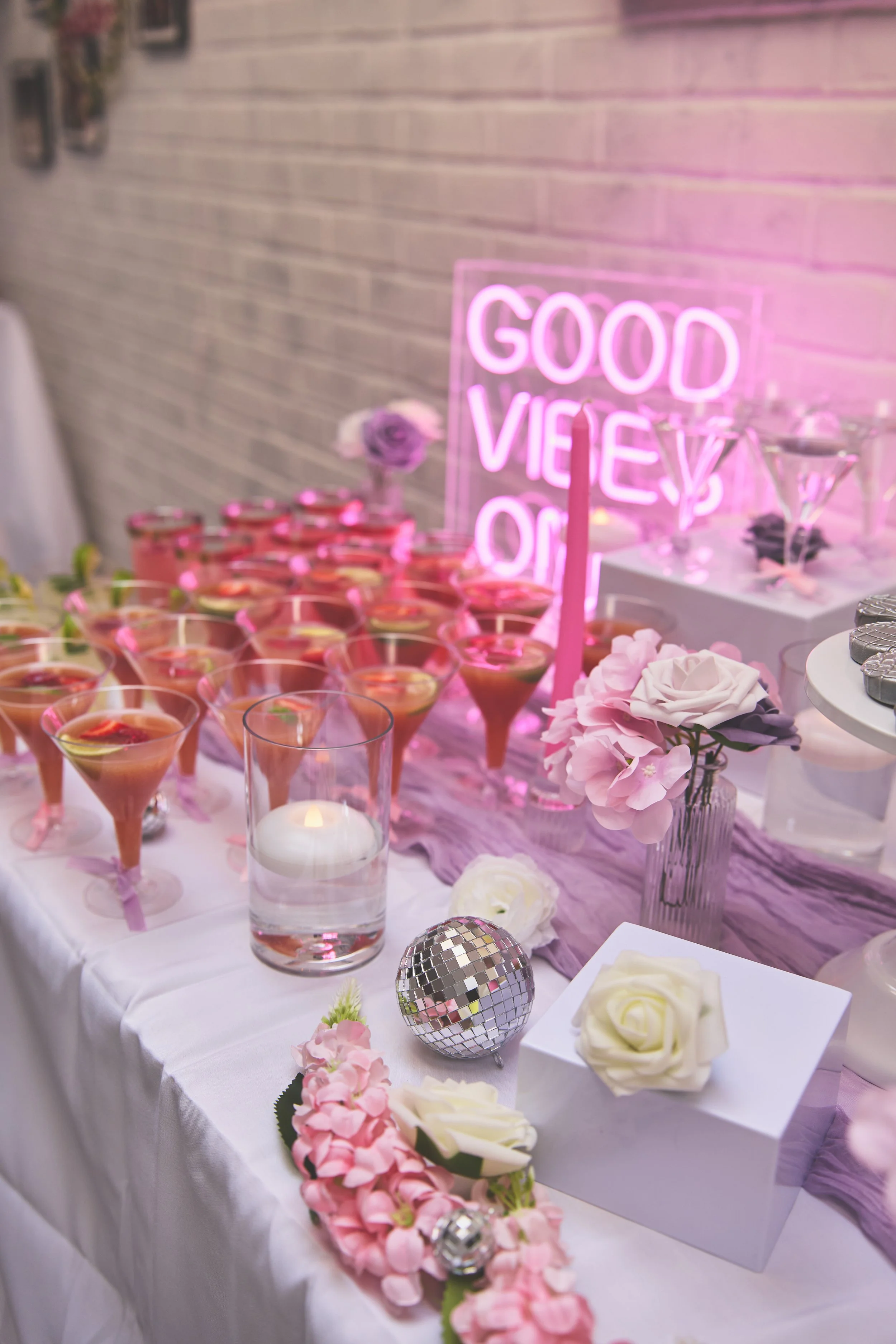 Decorative table with pink and white flowers, pink cocktails, candles, a disco ball, and a pink neon sign that reads 'Good Vibes Only' at a celebration or party.
