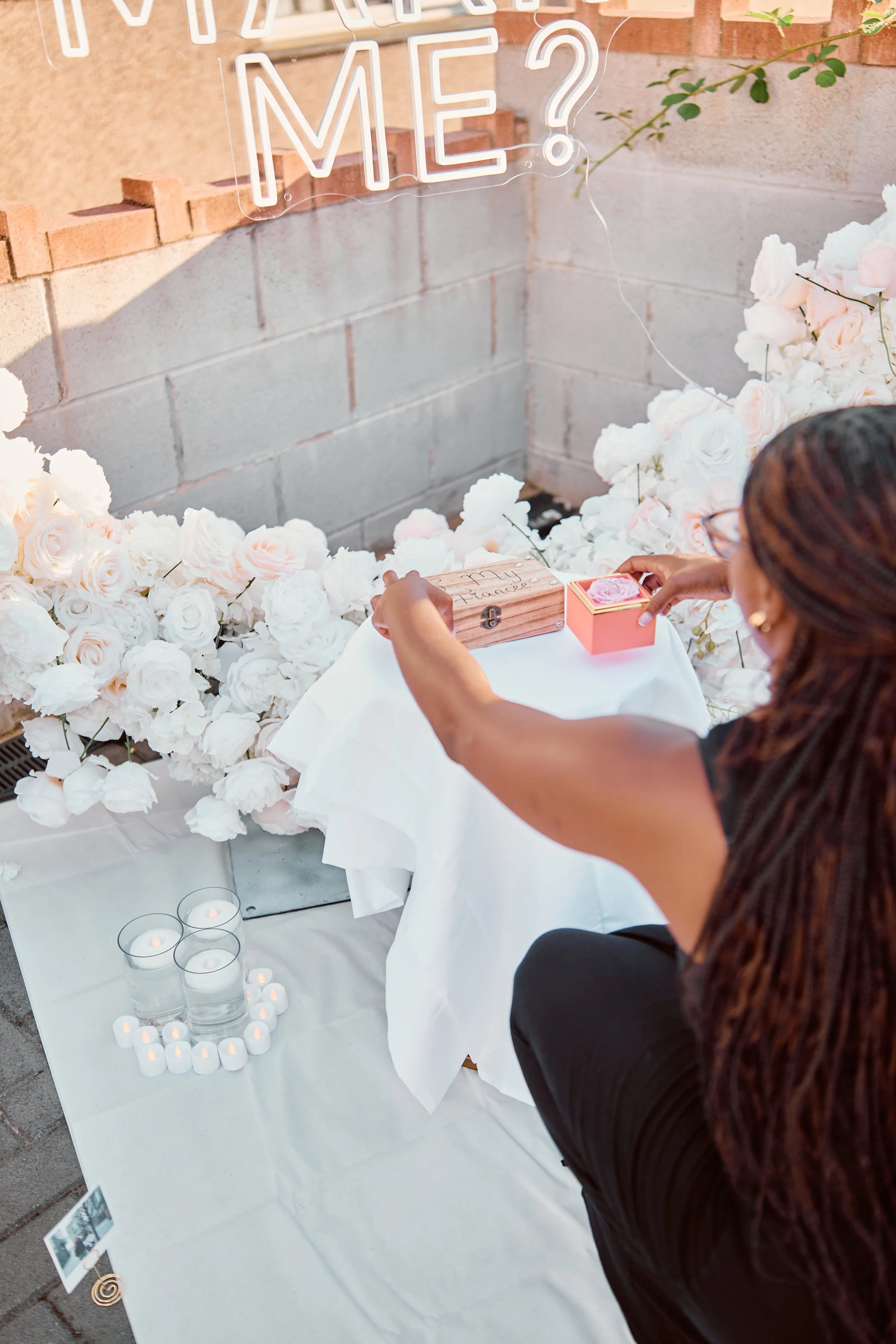 A woman is reaching to place a pink flower into a small pink box, atop a white table decorated with white roses, candles, and a wooden box with writing, against a brick wall with a glowing neon sign that reads 'Will you marry me?'.