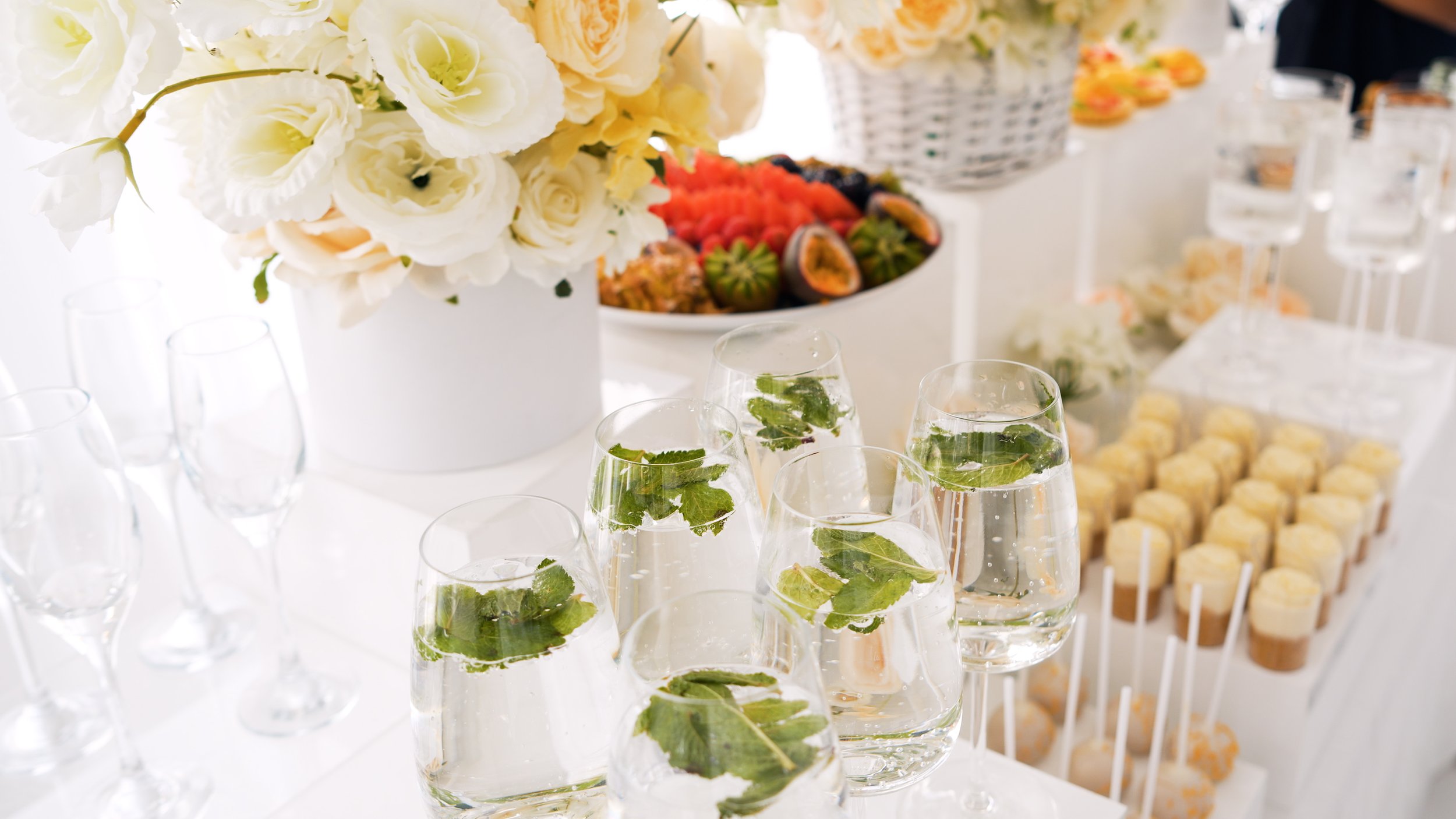 Table setting with a floral centerpiece, champagne flutes with mint leaves, a platter of sliced fruits, and mini desserts on sticks.