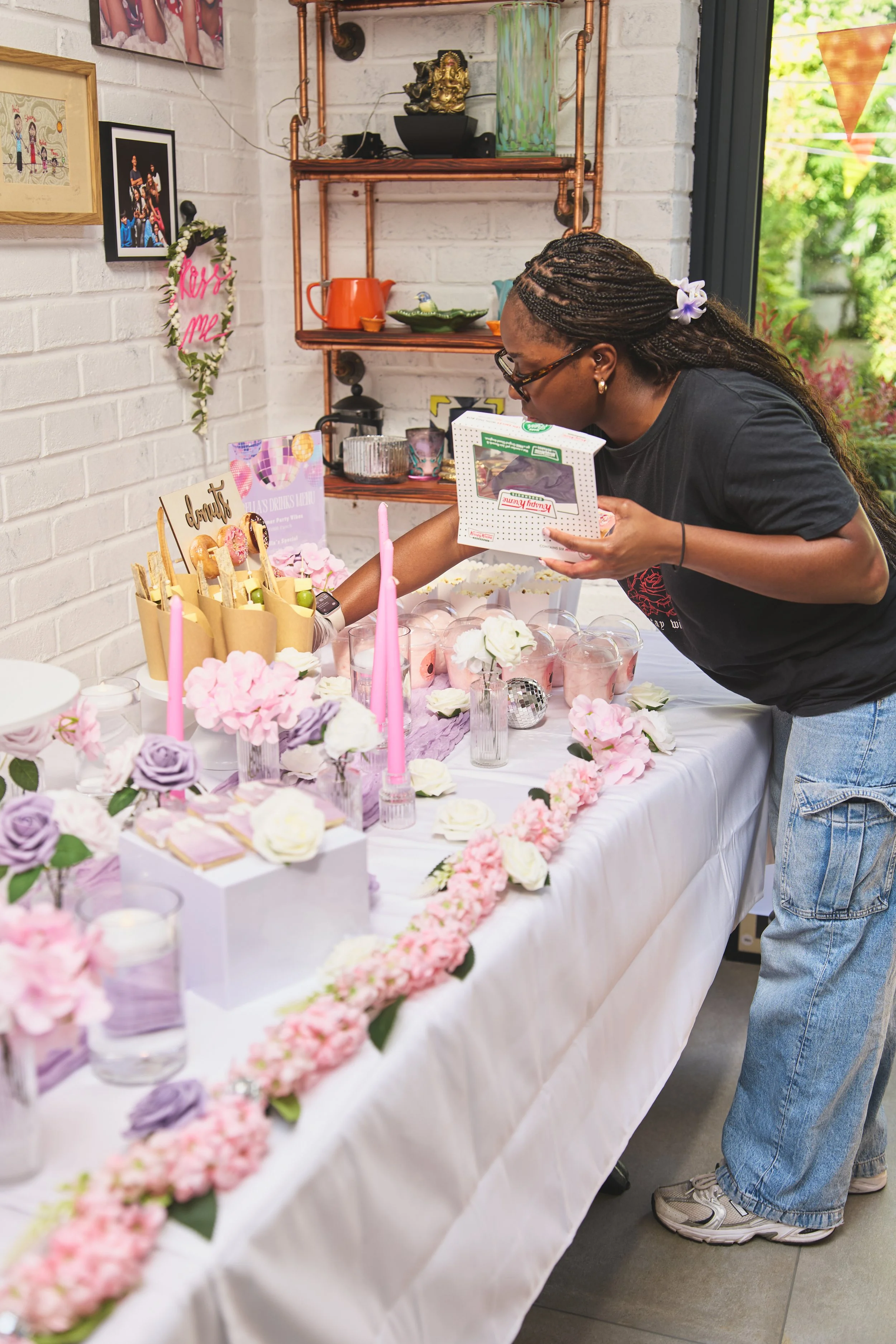 A woman with braided hair in glasses leaning over a decorated party table, arranging items for a celebration. The table is adorned with pink, purple, and white flowers, candles, and treats. In the background, decorative shelves and framed pictures are visible, as well as a window showing greenery outside.
