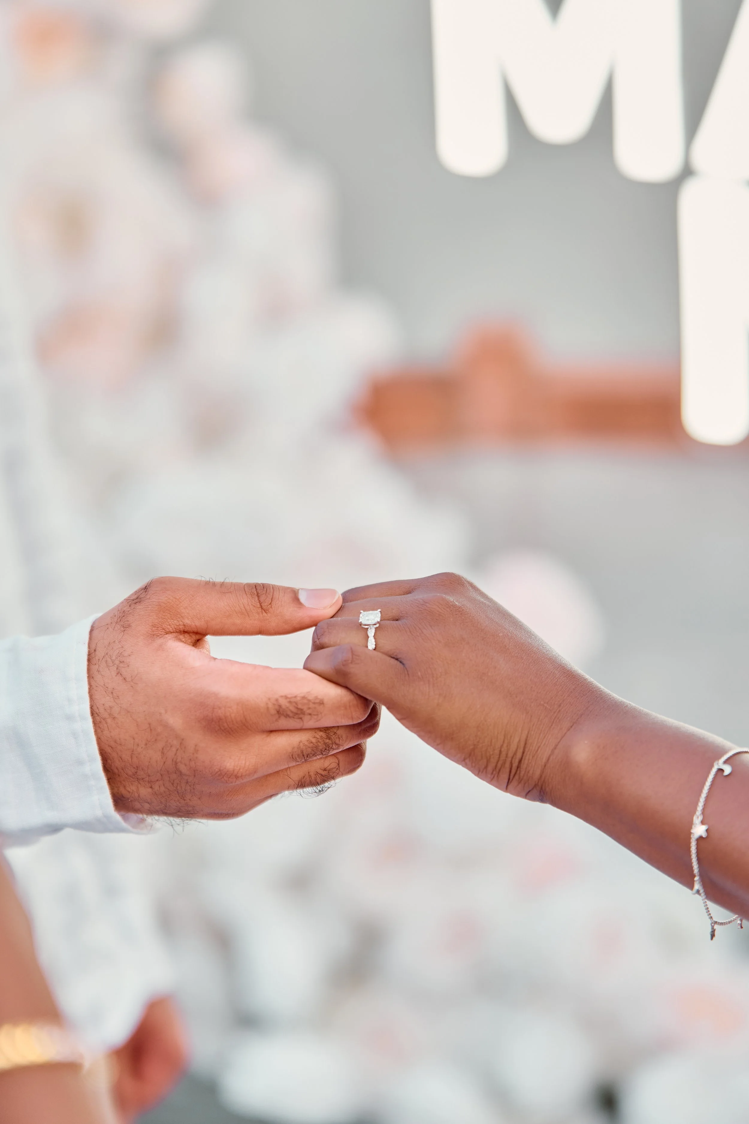 A man places a ring on a woman's finger during a wedding ceremony, with a blurred background of wedding decorations.