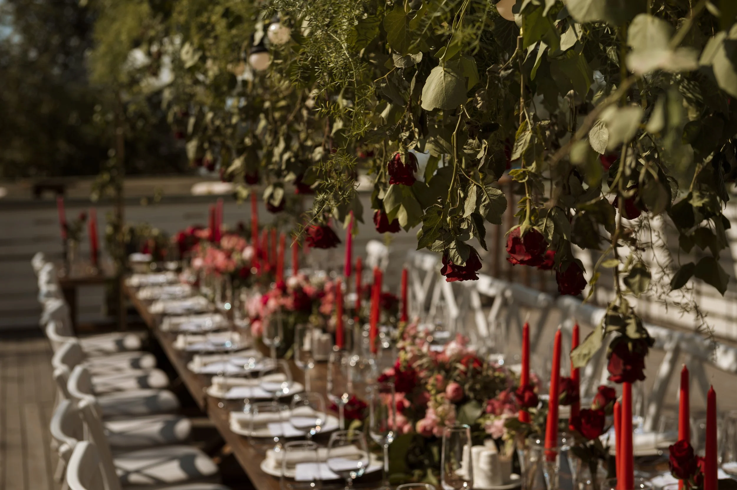 A long dining table set for an event with floral centerpieces and red candles, decorated with hanging green foliage and flowers.