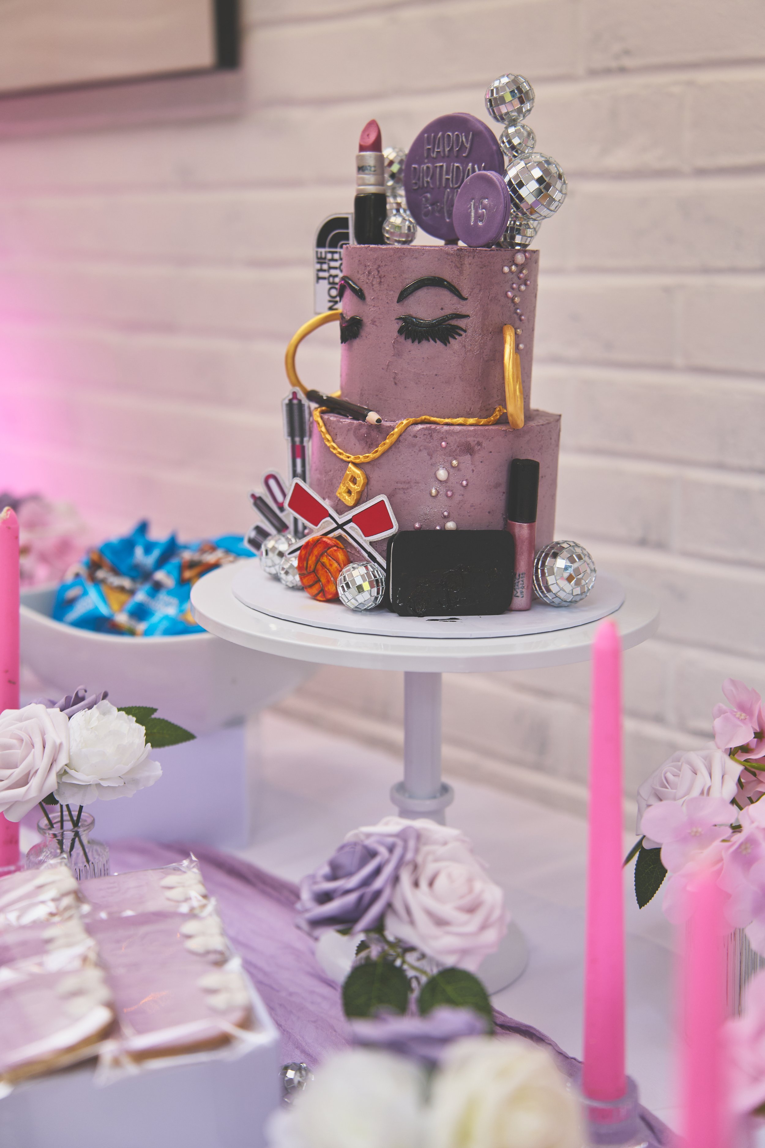 purple birthday cake on stand with candles and flowers around it