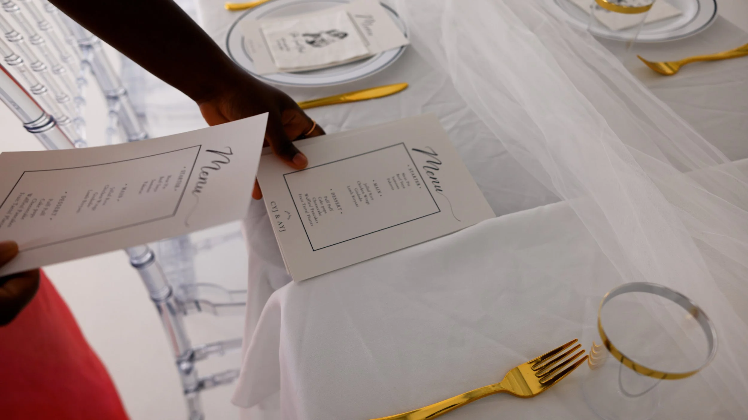 A person holding a restaurant menu over a white tablecloth, with gold cutlery and clear glasses nearby.