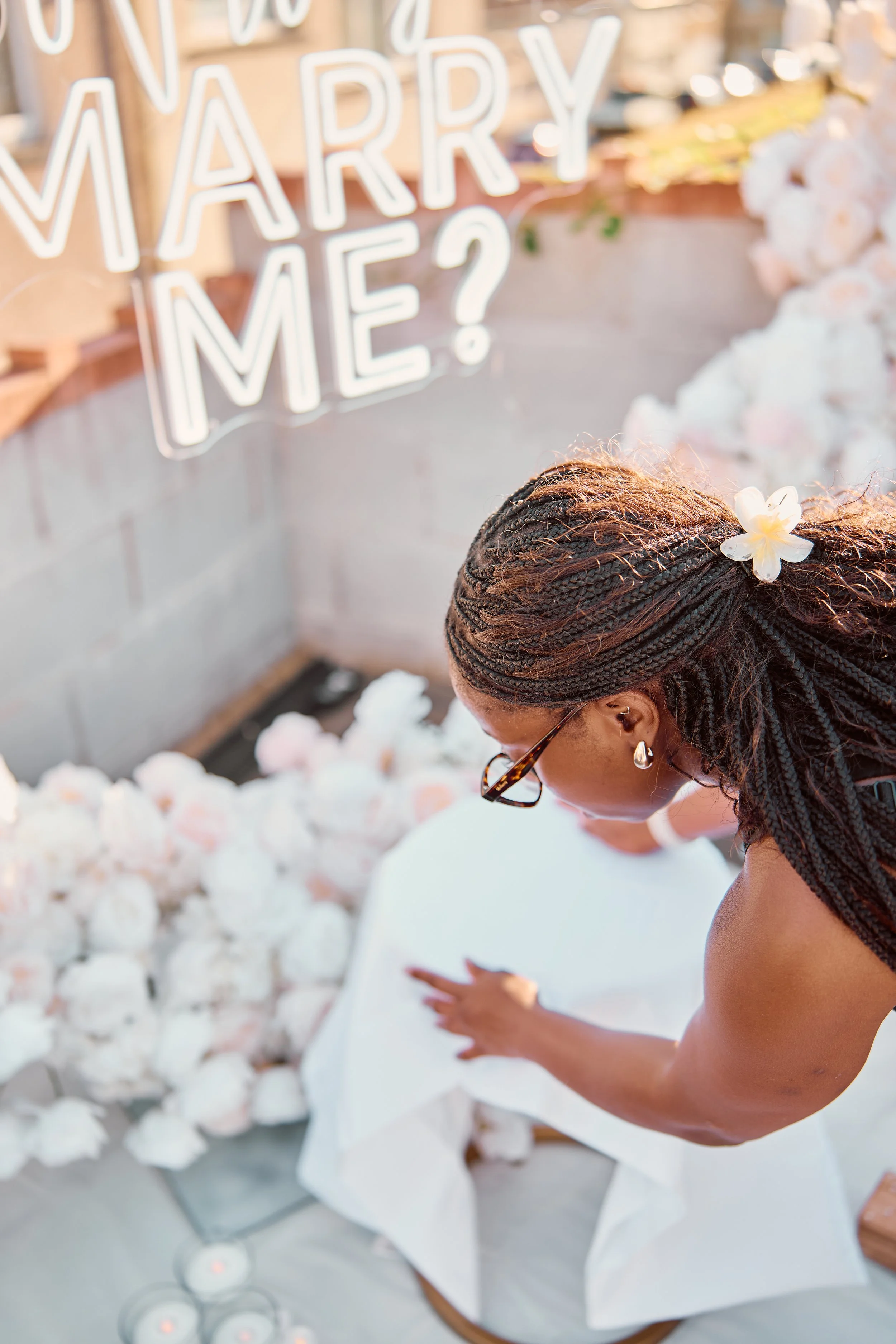 A woman with braided hair and glasses arranging white flowers on a table in front of a floral backdrop with a neon sign that reads 'Will You Marry Me?'
