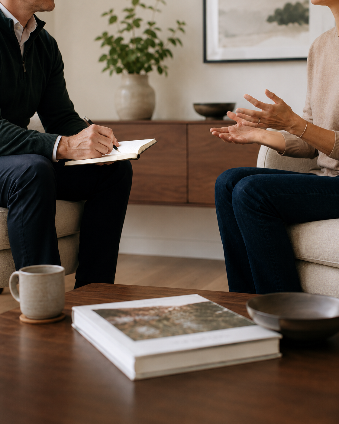 A therapist and client sitting in a cozy living room during a therapy session, with a wooden table in front holding a book, a cup, and a bowl. The therapist is taking notes, and the client is gesturing with hands.