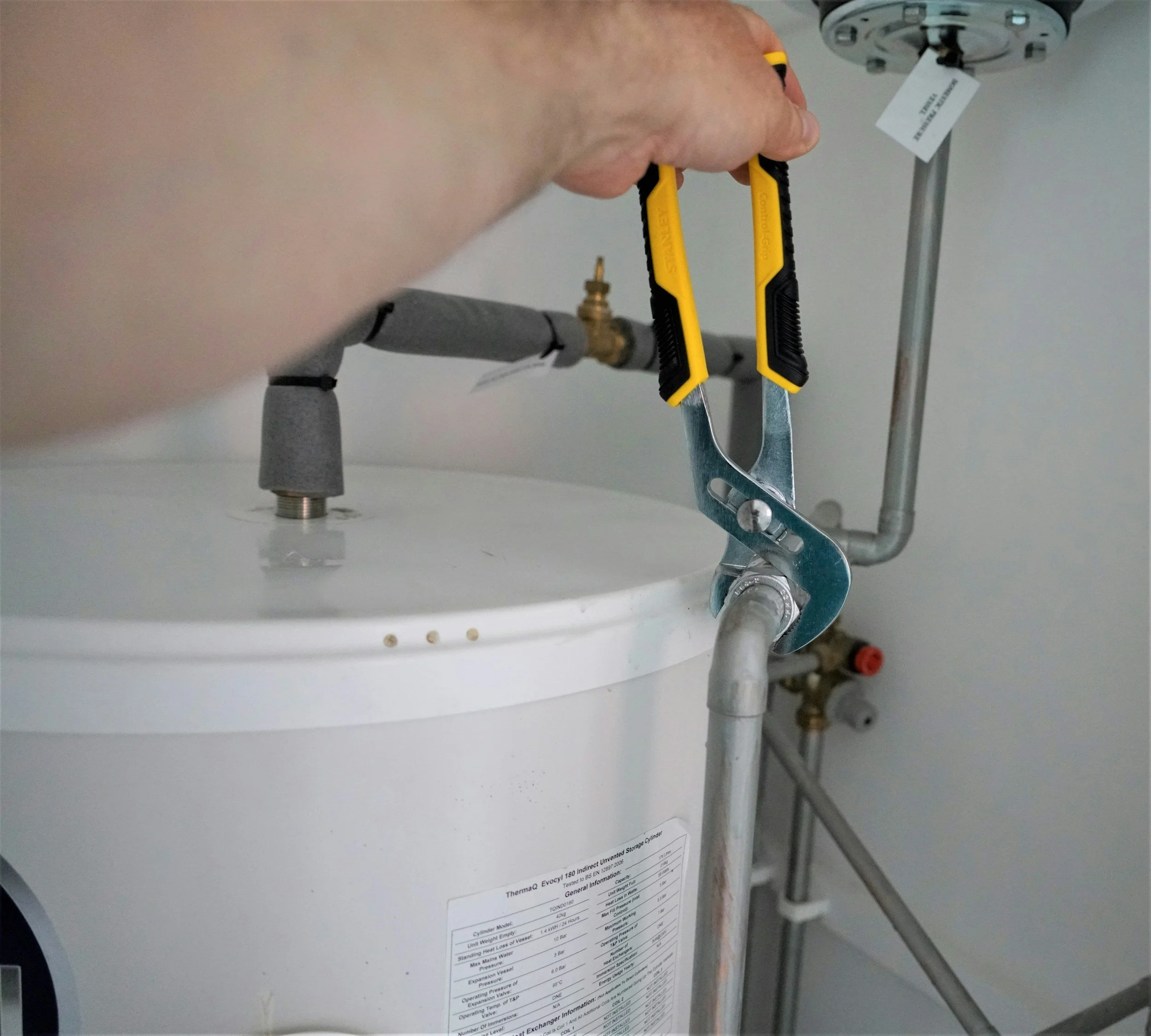 A person is using a wrench to tighten or loosen a fitting on a pipe under a sink. There are pipes and plumbing components visible, with a label or label information attached to the sink assembly.