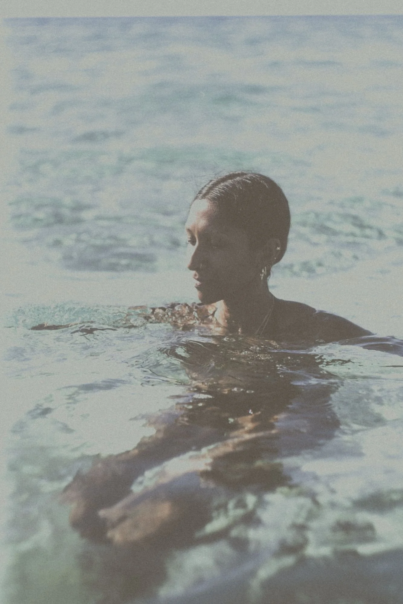 Nia with wet hair and earrings, partially submerged in water at the beach, facing sideways with a calm expression.
