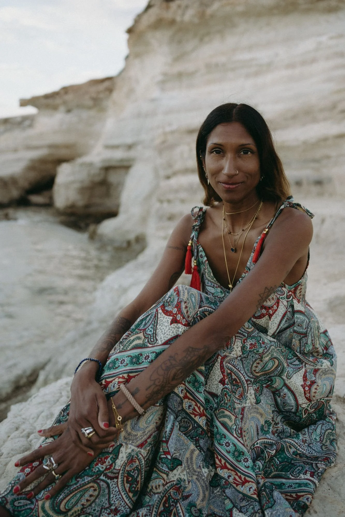Nia sitting on sandy rocks by the beach, wearing a colorful, patterned dress, with tattoos on her arms, and jewelry including necklaces, rings, and bracelets, with a rocky cliff in the background.