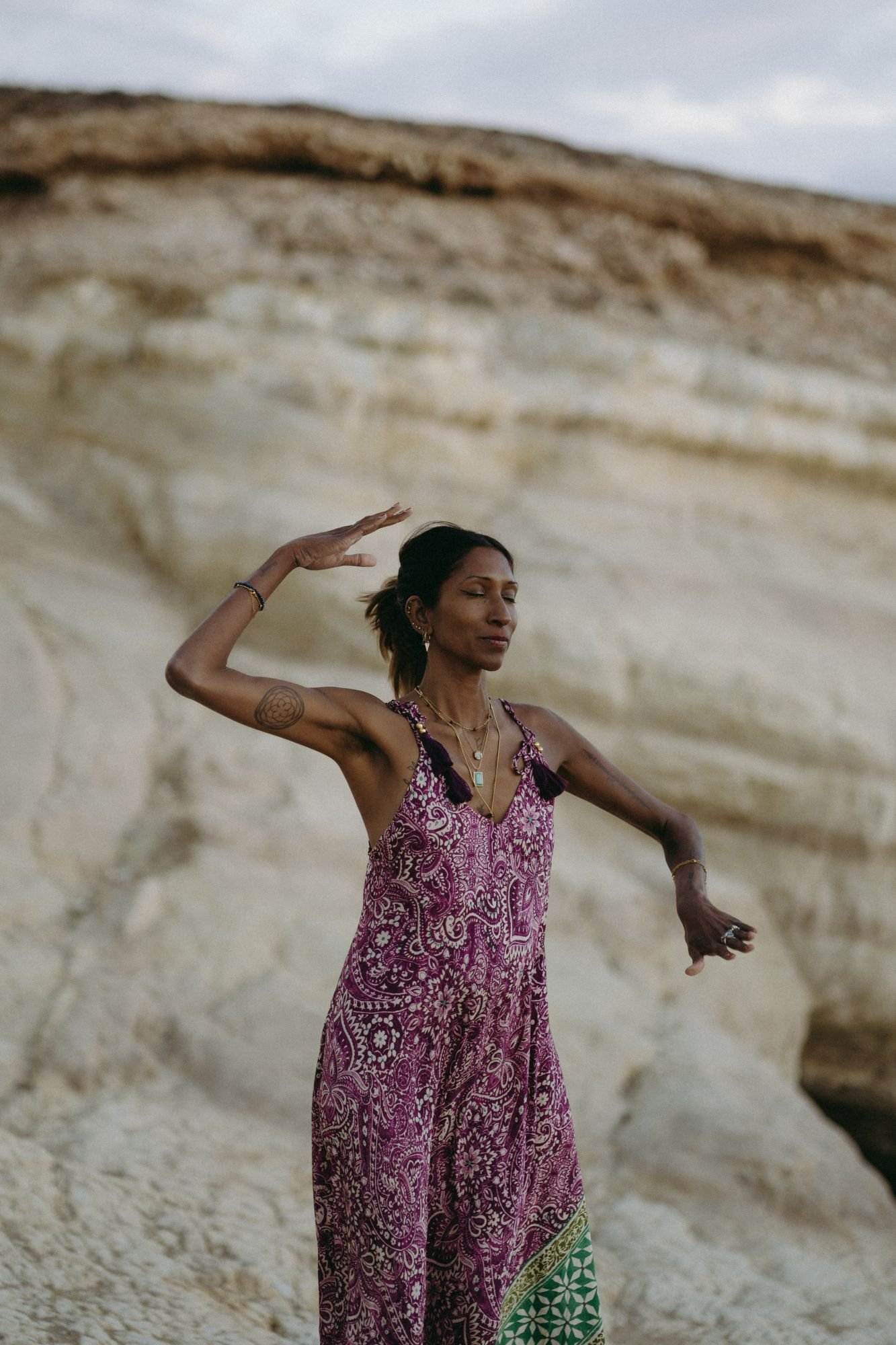 Nia with dark hair and tattoos, wearing a colorful patterned dress, stands with her eyes closed on a rocky outdoor landscape, engaged in light language.