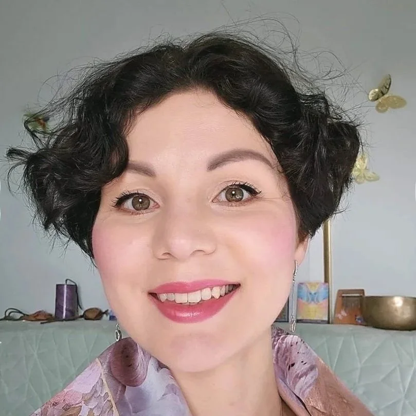 Darya, A woman with short, curly dark hair smiling, wearing earrings and a pastel-colored scarf, in a room with decorative items, including a large bowl and framed pictures.