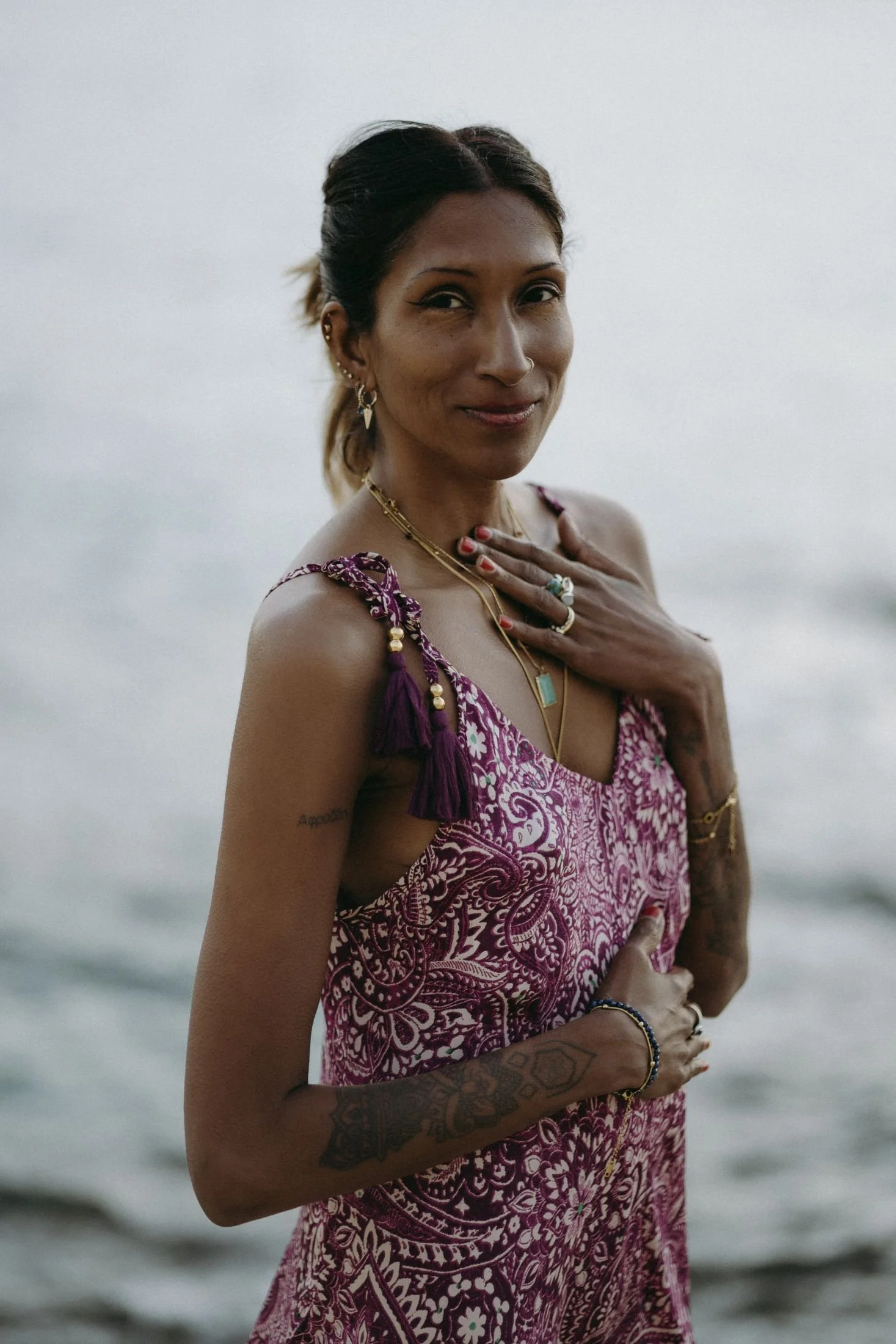 Nia in a purple patterned dress standing outdoors by the water at sunset, with jewelry and tattoos.