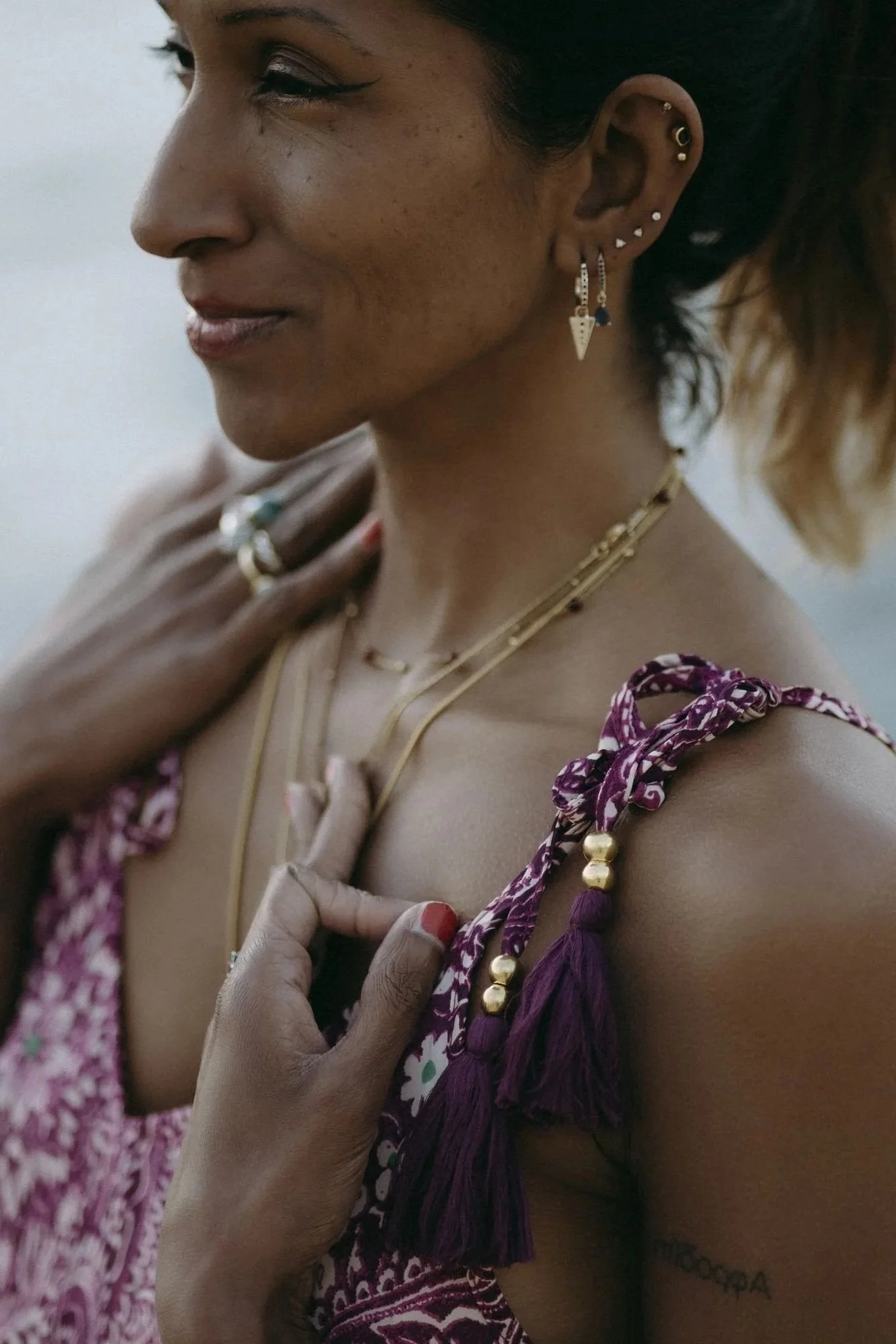 A woman wearing gold earrings and layered gold necklaces, with a colorful, patterned top that has purple tassels on the straps. She has a tattoo on her arm.