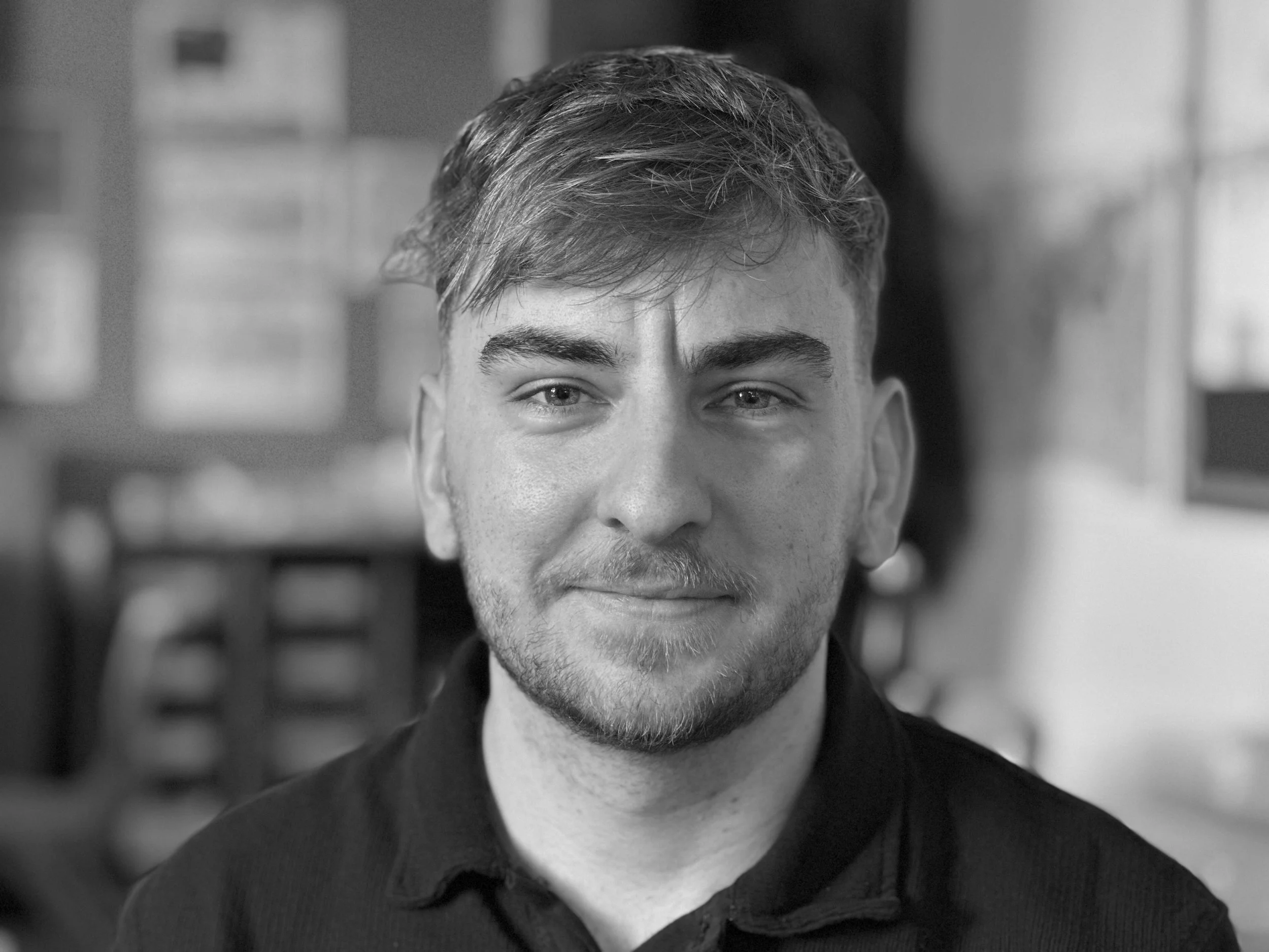 A black and white photo of a young man with short, slightly wavy hair, slight beard, and a minor smile, wearing a dark collared shirt, indoors with shelves and blurred objects in the background.