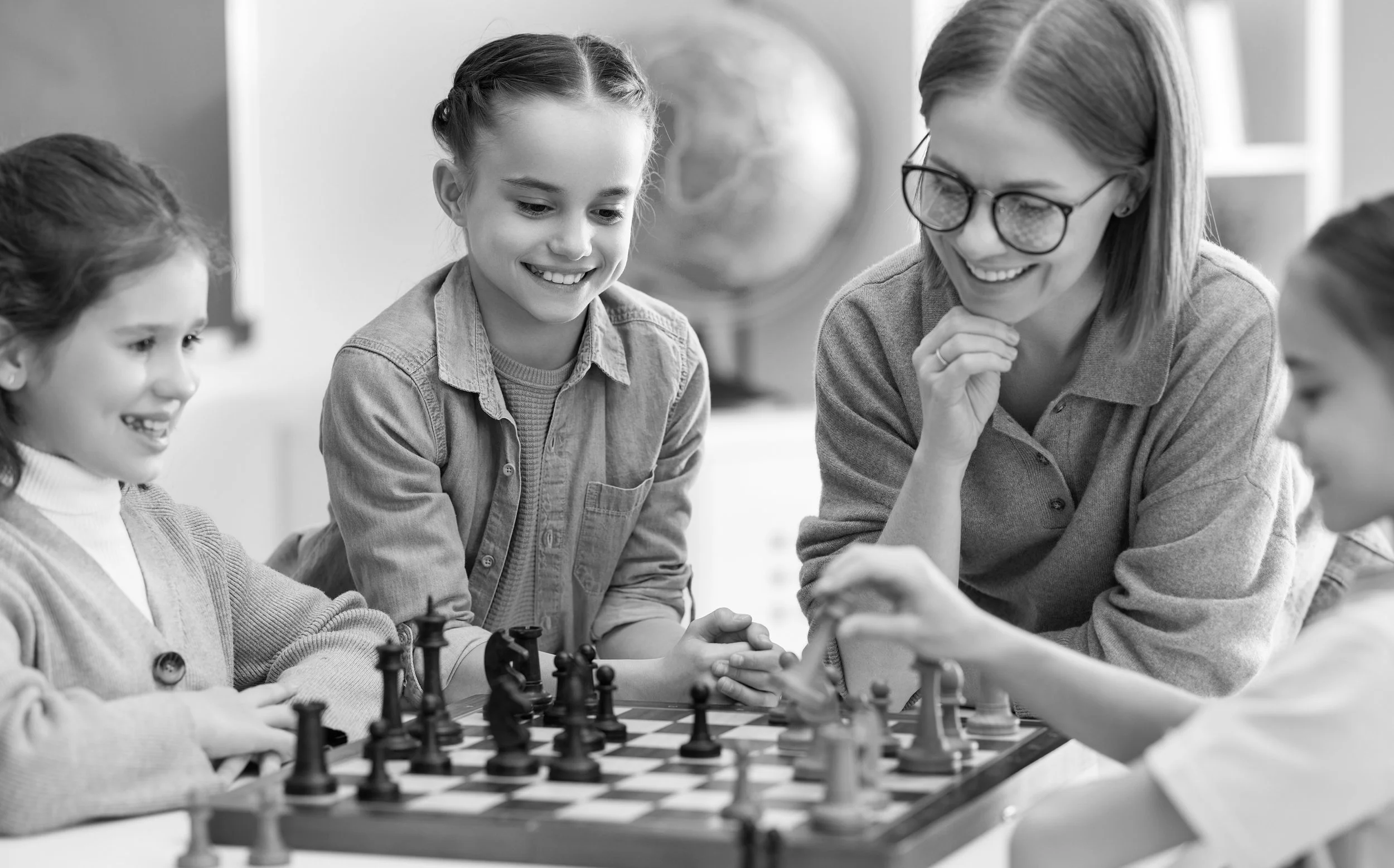 A group of children and an adult girl playing chess. They are sitting at a table with a chessboard, smiling and engaging in the game.