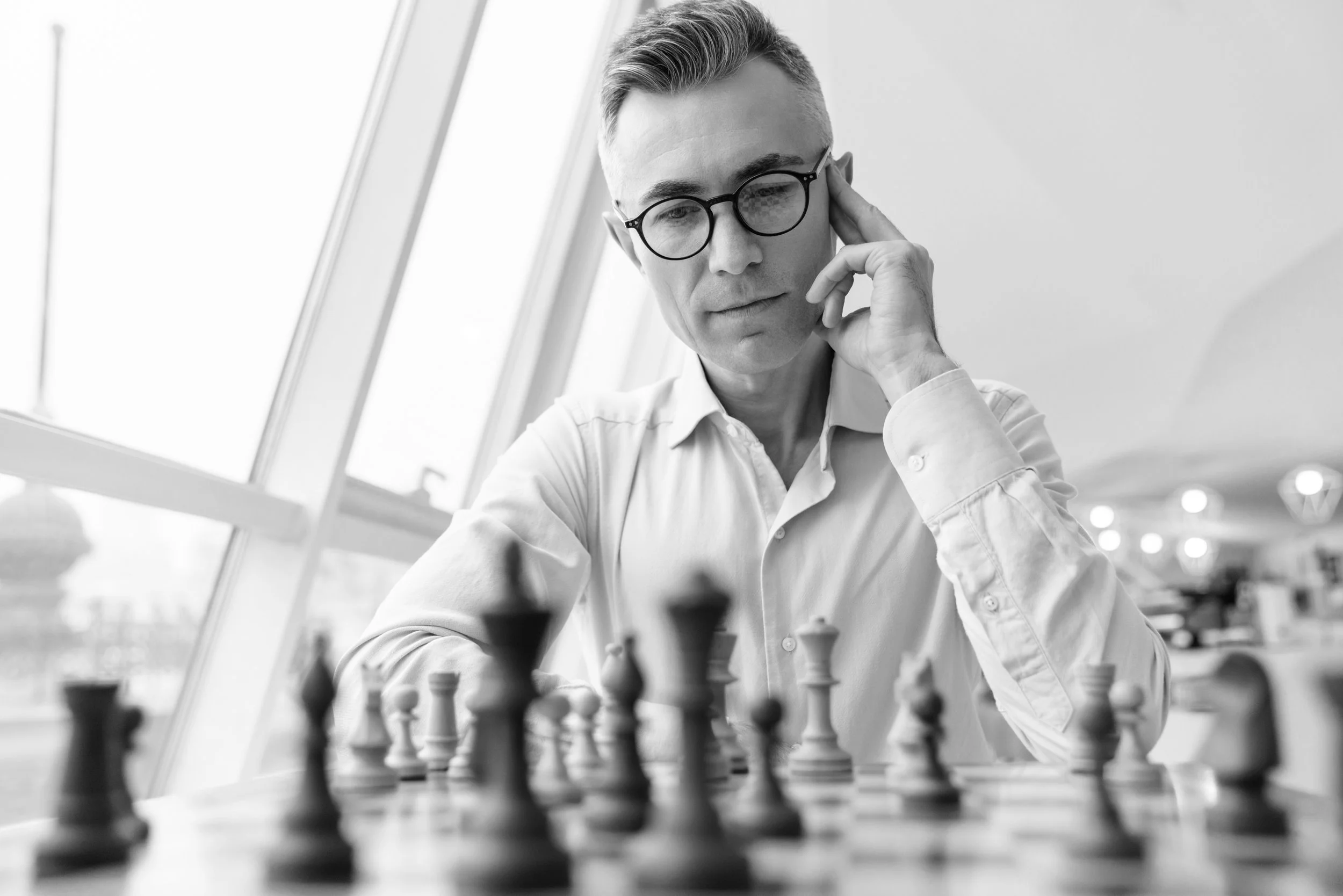 A man with glasses and a white shirt playing chess in a well-lit indoor space.