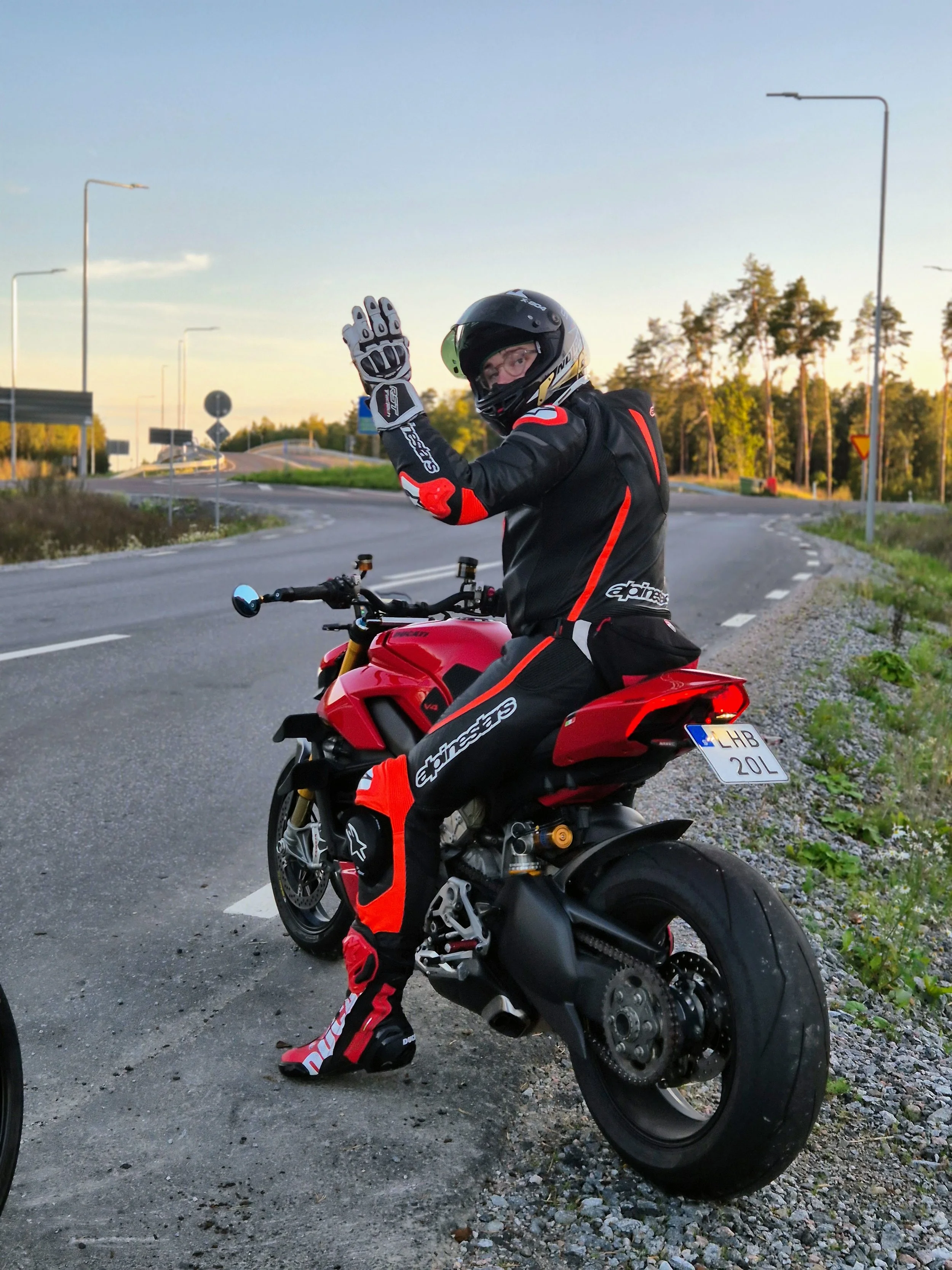 A person wearing a motorcycle racing suit, helmet, and gloves sitting on a red motorcycle, waving at the camera, parked by the side of a road with trees and streetlights in the background during sunset.