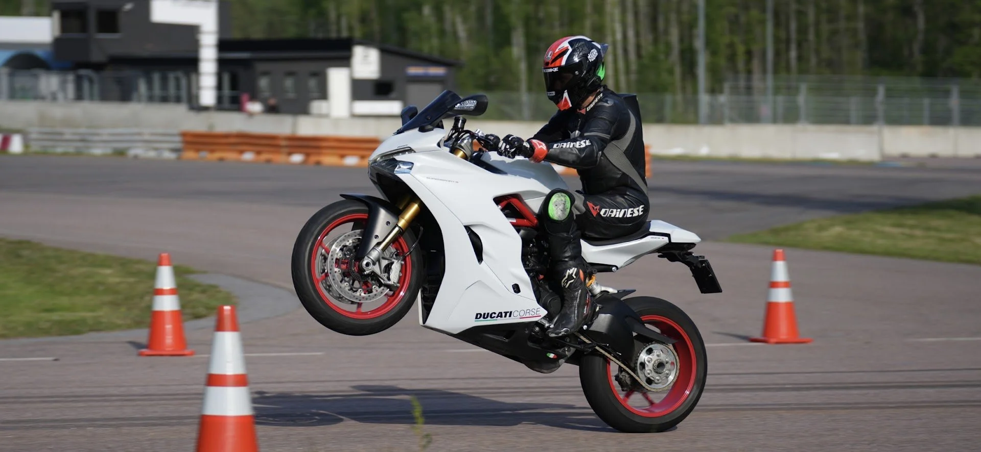 Motorcyclist performing a wheelie on a white Ducati motorcycle during a training or racing session, wearing a black full-body suit and helmet, on a paved track marked with orange safety cones.