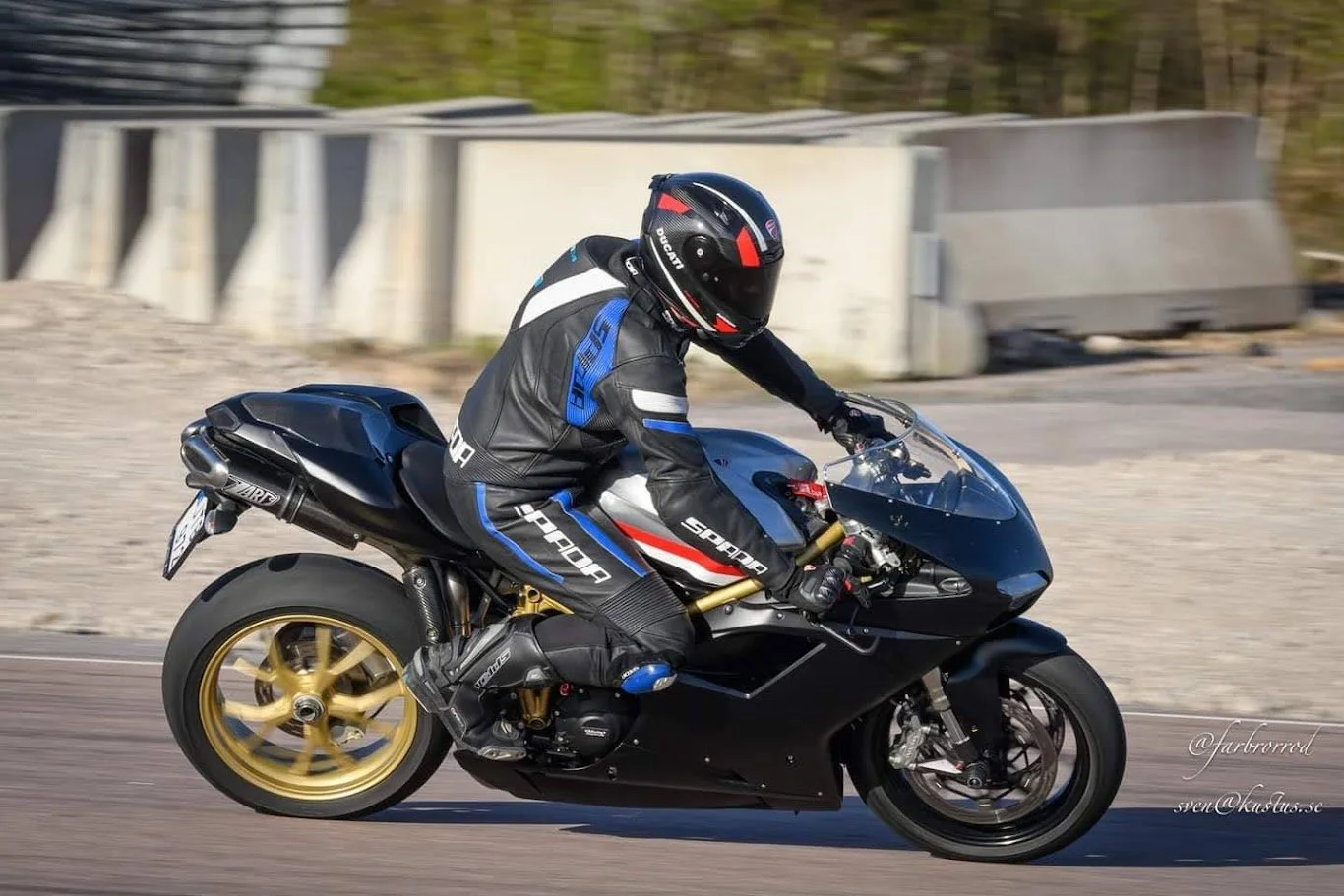 A motorcyclist wearing a black, blue, and white racing suit and a black helmet rides a black sport motorcycle on a paved road.