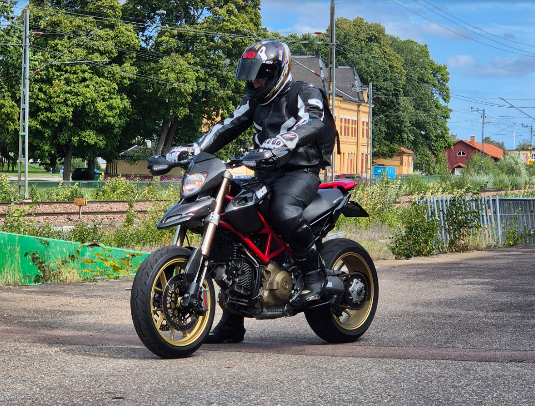 Motorcyclist in black leather gear and helmet riding a black and red motorcycle on a street with trees and buildings in the background.