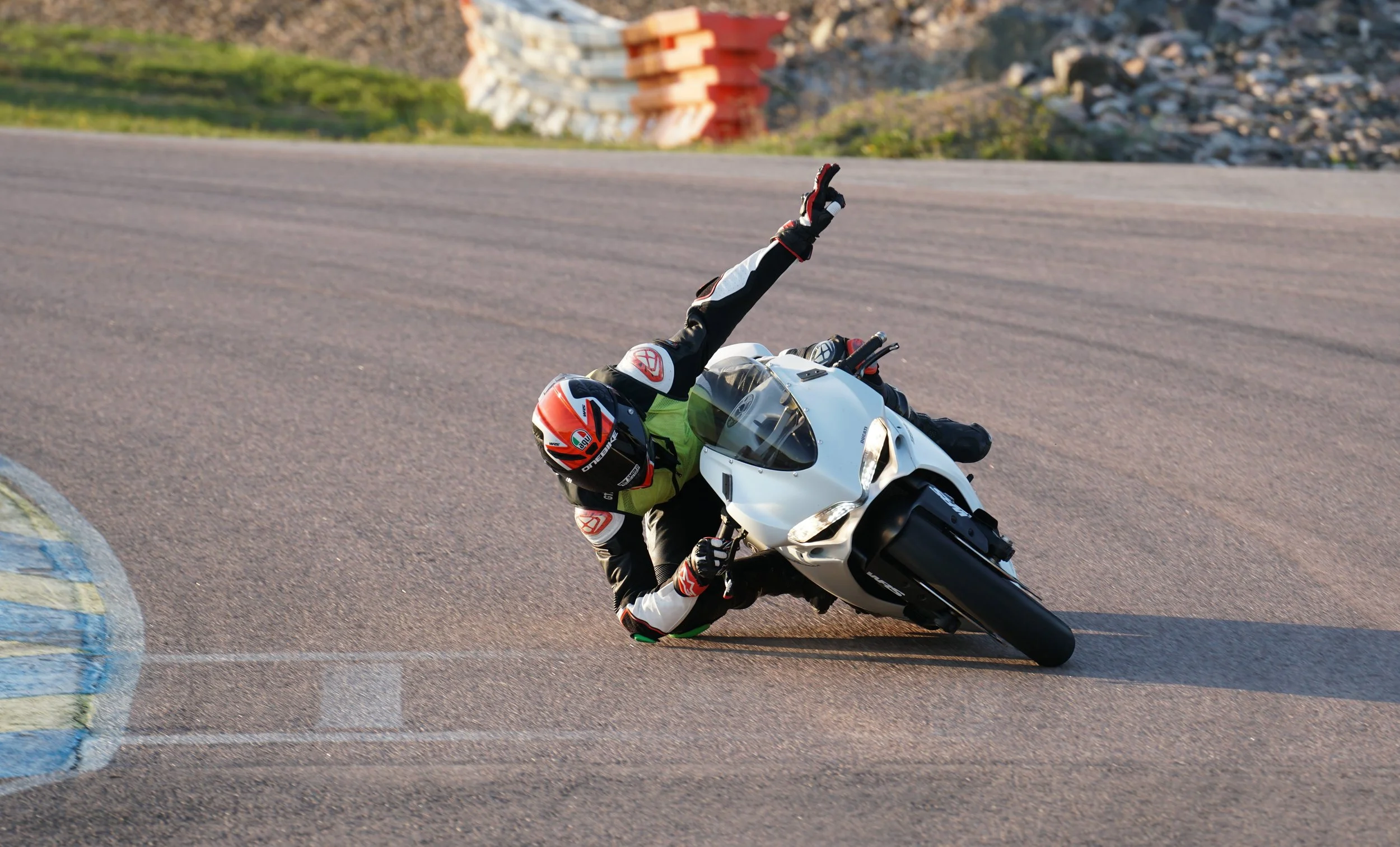 A motorcyclist in racing gear leans sharply into a turn on a racetrack, with the motorcycle tilting at a steep angle. The rider's arm is extended, making a gesture with two fingers raised. The background shows a blurred section of the track and some 