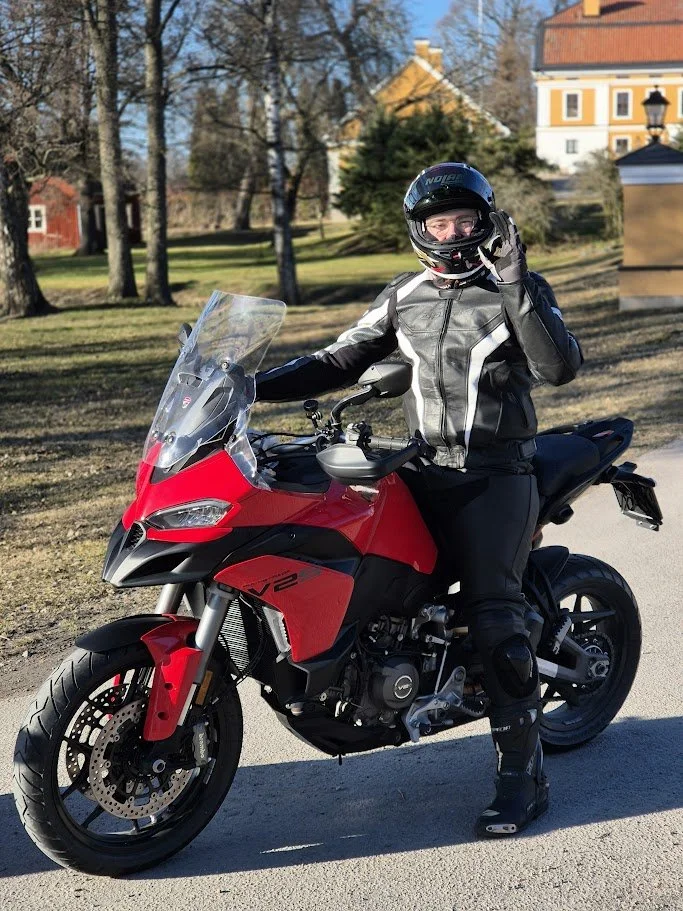 A person wearing a motorcycle helmet, jacket, and protective gear standing next to a red motorcycle on a sunny day with trees and houses in the background.