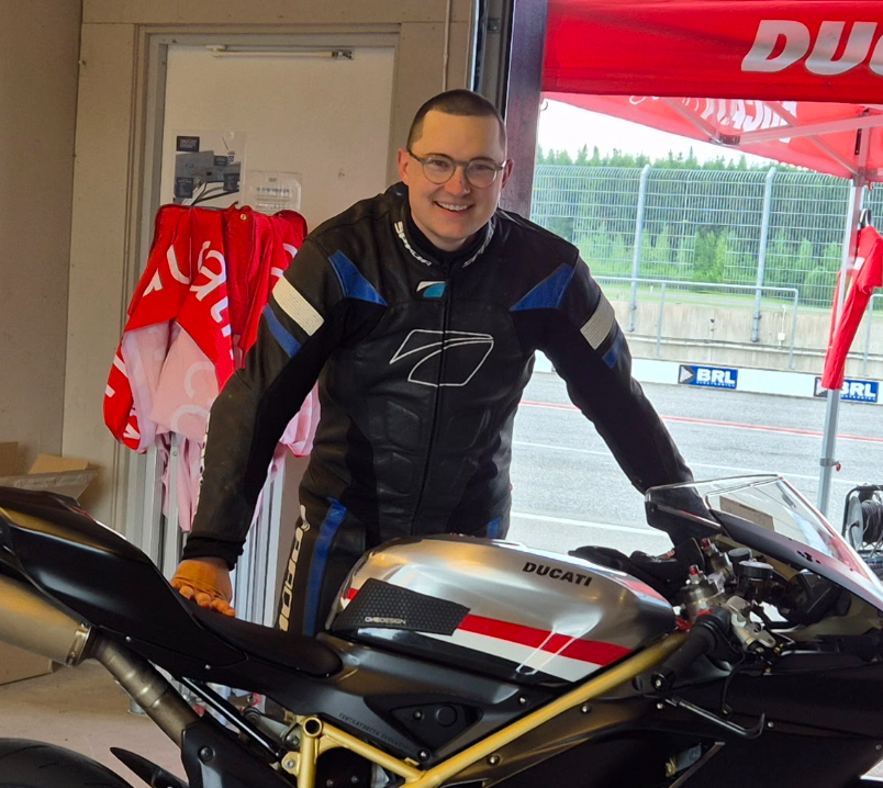 A smiling man in a racing suit standing next to a Ducati motorcycle inside a motorsport garage.