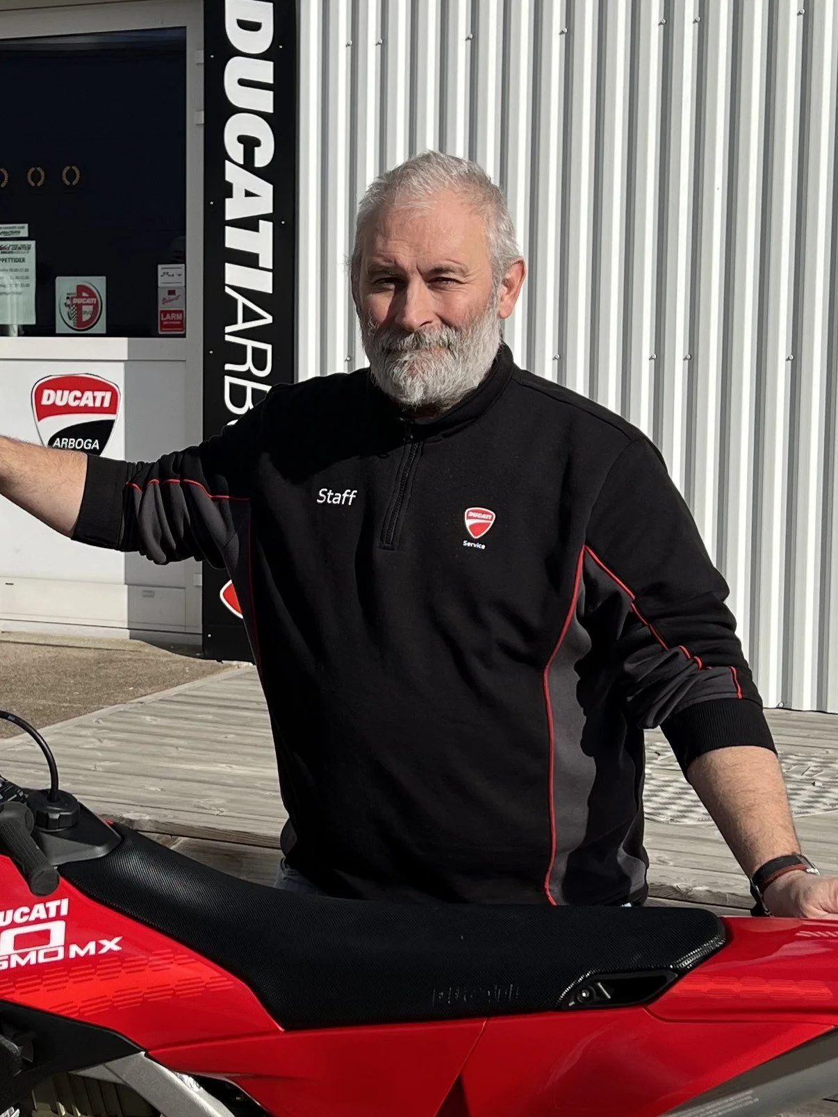 A man with gray hair and beard standing next to a red Ducati motorcycle outside a Ducati service shop, wearing a black Ducati staff jacket.