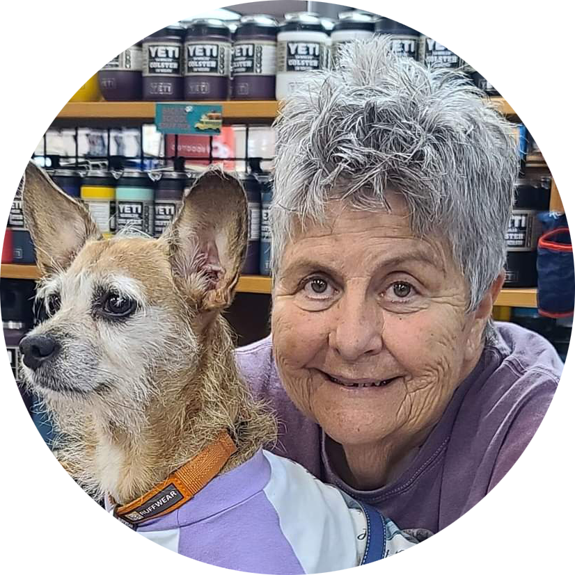 An elderly woman smiling with a scruffy tan and white dog with large ears in a store aisle lined with jars of paint in the background.