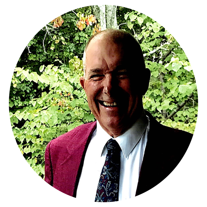 Smiling man in a suit and tie, outdoors with green leafy background.
