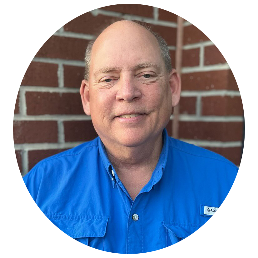 A middle-aged man with a slight smile, wearing a blue Columbia outdoor shirt, standing in front of a red brick wall.