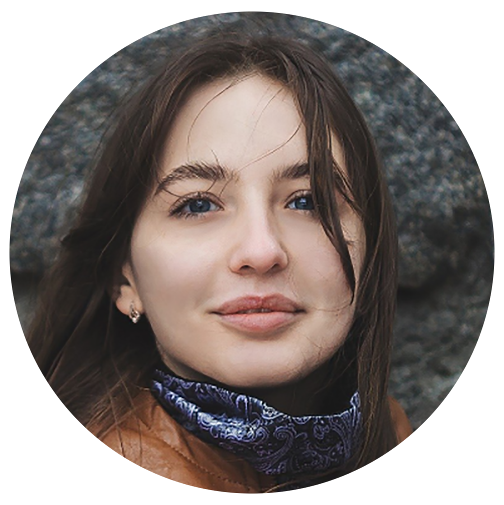 Close-up of a young woman with brown hair, blue eyes, and a slight smile, outdoors with rocks in the background.