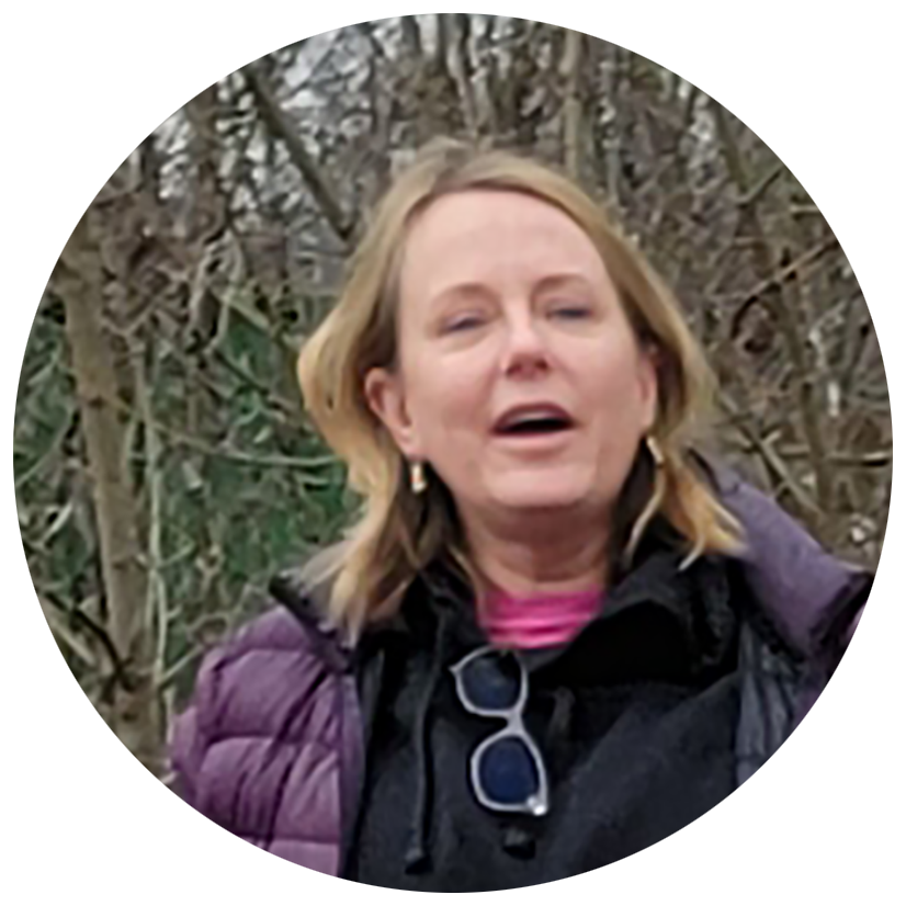 A woman with blonde hair wearing a purple jacket and pink shirt outdoors among leafless trees.