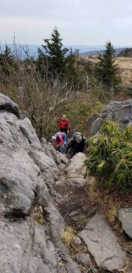 Three hikers climbing a rocky mountain trail surrounded by trees and shrubs.
