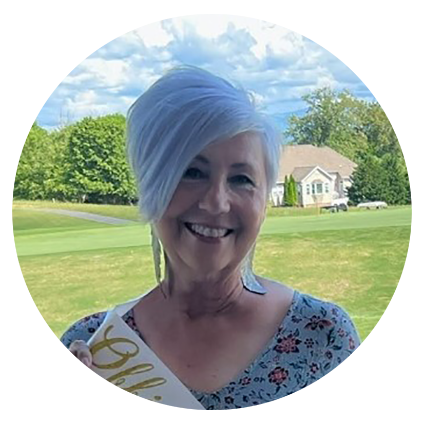 Smiling woman with short, asymmetrical white and light blue hair, holding a white umbrella with gold lettering, outdoors in a green park with trees and houses in the background, under a partly cloudy sky.