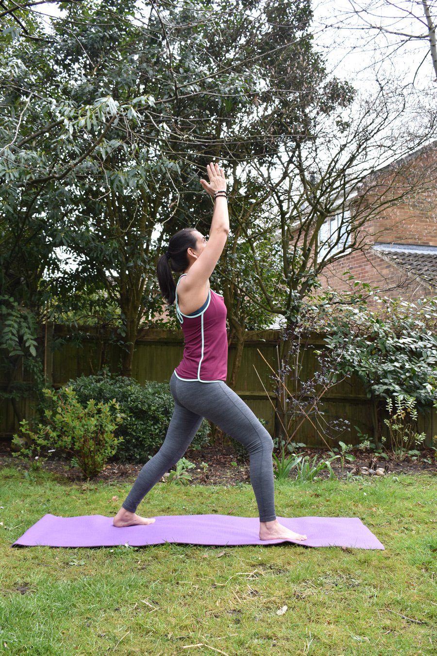 Sophia Armstrong practicing yoga, standing on a purple yoga mat, raising her arms overhead, surrounded by trees and bushes.